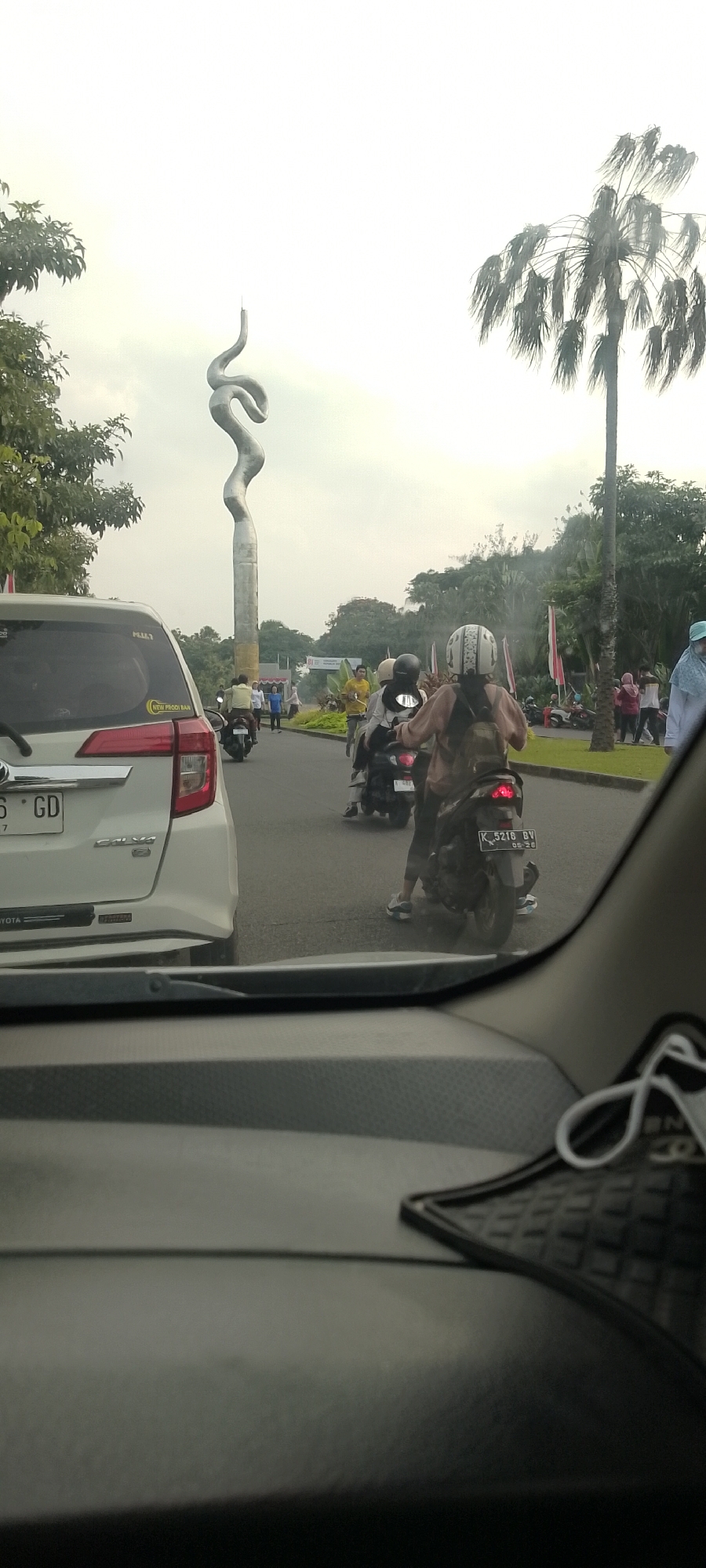 In Bacin, Indonesia, a traffic scene unfolds with a prominent, tall, serpentine sculpture dominating the background against an overcast sky. The sculpture is a metallic, silver-grey structure that twists and turns upwards. To the right of the sculpture, a tall palm tree with fronds extending outwards is visible. In the midground, a street is lined with trees and grass. Motorcyclists are a significant element, with several riding in the same direction. One motorcycle in the foreground has a rider wearing a light brown jacket and a white helmet, facing away from the camera. Another motorcycle behind it has a rider with a black helmet. A white car is also present in the foreground, with its rear visible to the left. License plates are partially visible on the white car and a motorcycle. People are scattered throughout the scene, some walking and some on motorcycles. Flags, possibly red and white, are displayed in the midground. The image appears to be taken from inside another vehicle, as the dashboard and windshield wipers are visible in the bottom portion of the frame.