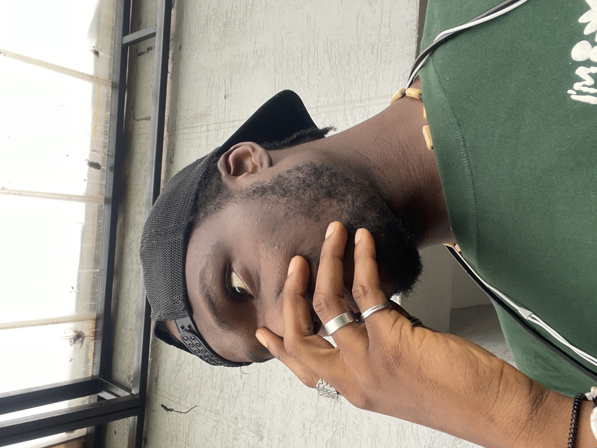 A man wearing a black baseball cap and a green t-shirt is shown from the chest up, positioned outdoors in front of a textured, light-colored wall. His right hand is raised, with his fingers covering his mouth and nose. He is looking to his left, away from the camera. He has a short beard and is wearing two silver rings on his right hand and a beaded necklace. The background includes a section of a structure with a translucent roof, supported by dark metal beams. The lighting suggests an overcast day. The scene is located in Abuja, Nigeria.