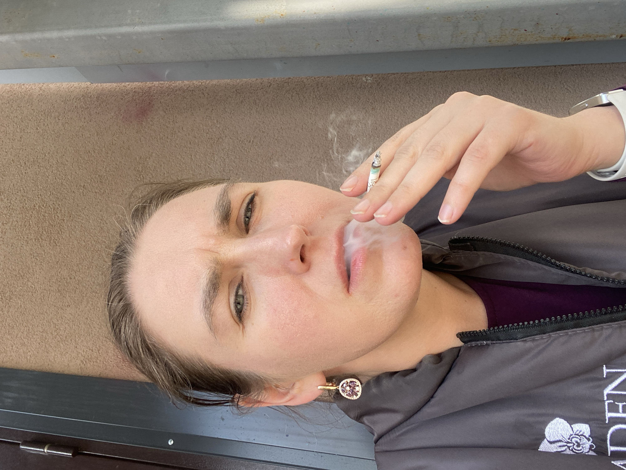 A woman is depicted in a close-up, angled selfie. She is holding a lit cigarette, with smoke emanating from her mouth. She is wearing a dark-colored zip-up jacket with a floral design and text, as well as a white wristwatch. She has light-colored skin, green eyes, and is wearing a gold earring with a decorative stone. The background includes a beige-colored wall with some imperfections and a metallic ledge. There is no information to conclude that the location is in the city of Yekaterinburg, Russia.