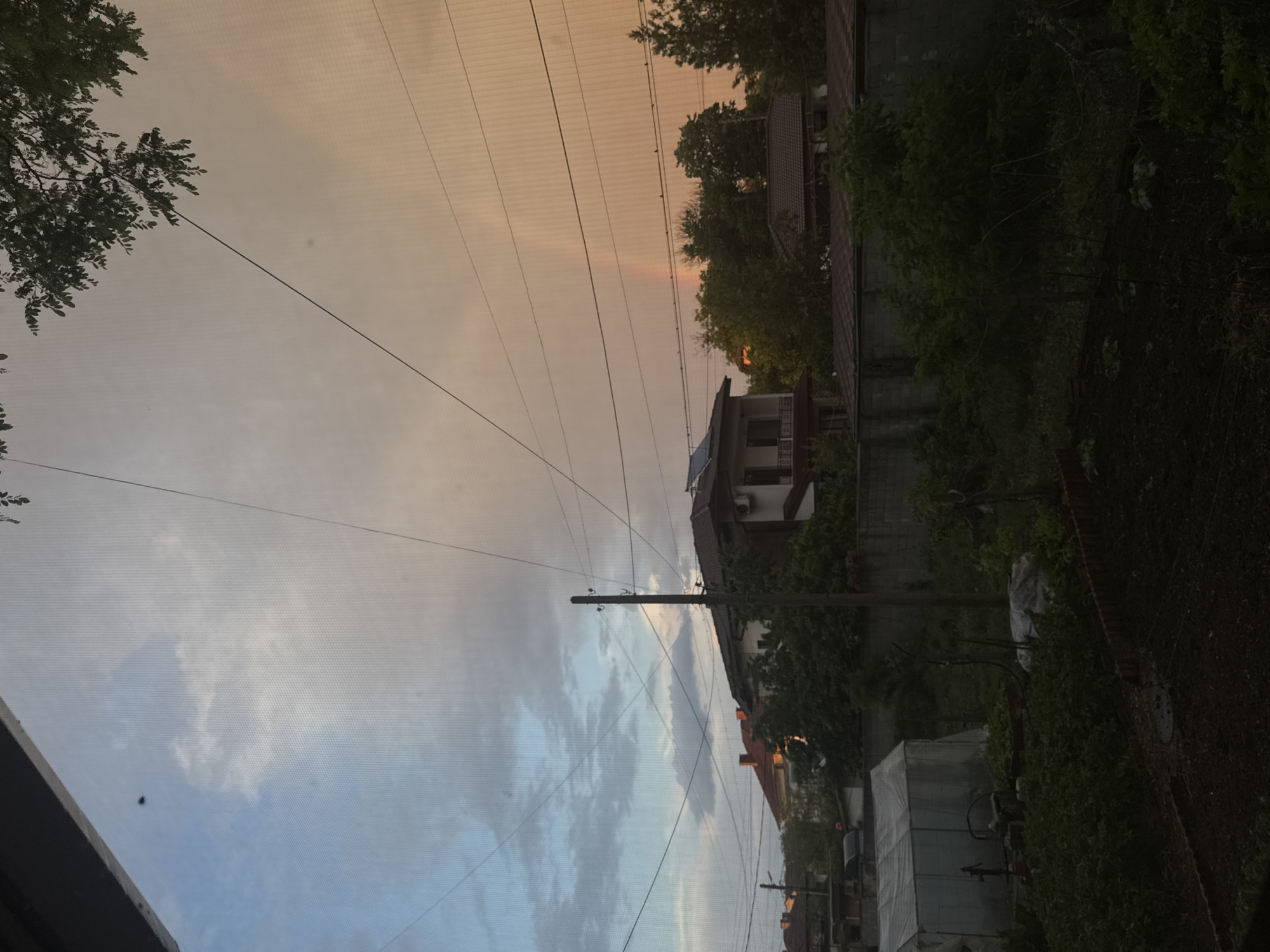The image depicts a residential scene in Elhovo, Bulgaria. A two-story house with a red roof and solar panels is visible, surrounded by greenery and a concrete fence. Utility poles and power lines dominate the sky, which transitions from blue with clouds to a light orange hue, possibly indicating sunrise or sunset. A portion of a mesh screen is visible in the foreground. Adjacent buildings and vegetation suggest an urban or suburban setting. A parked or covered vehicle can be seen on the right side. No people are visible in the image.