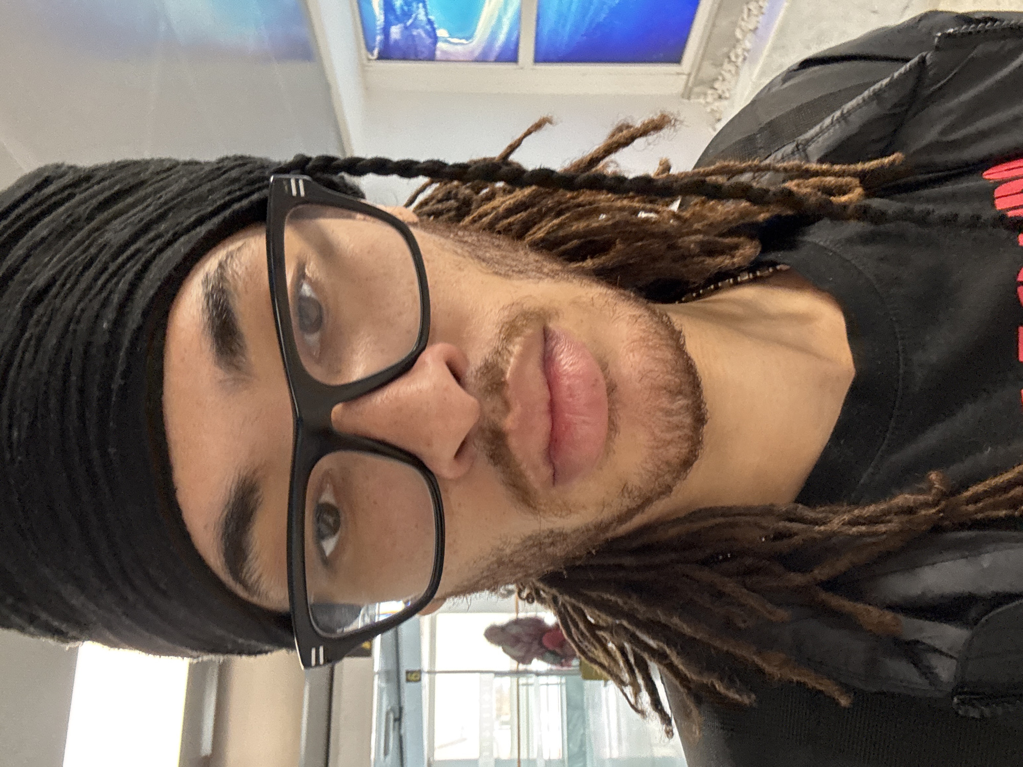 A close-up, tilted selfie shows a person with dreadlocks wearing a black head covering and black-rimmed glasses. The individual has facial hair and appears to be inside a building. The background is blurry, but a window with a blue-toned picture is visible. A partially visible black garment is on the subject's body. Details suggesting a location in San Bartolomé, Spain, are not present.