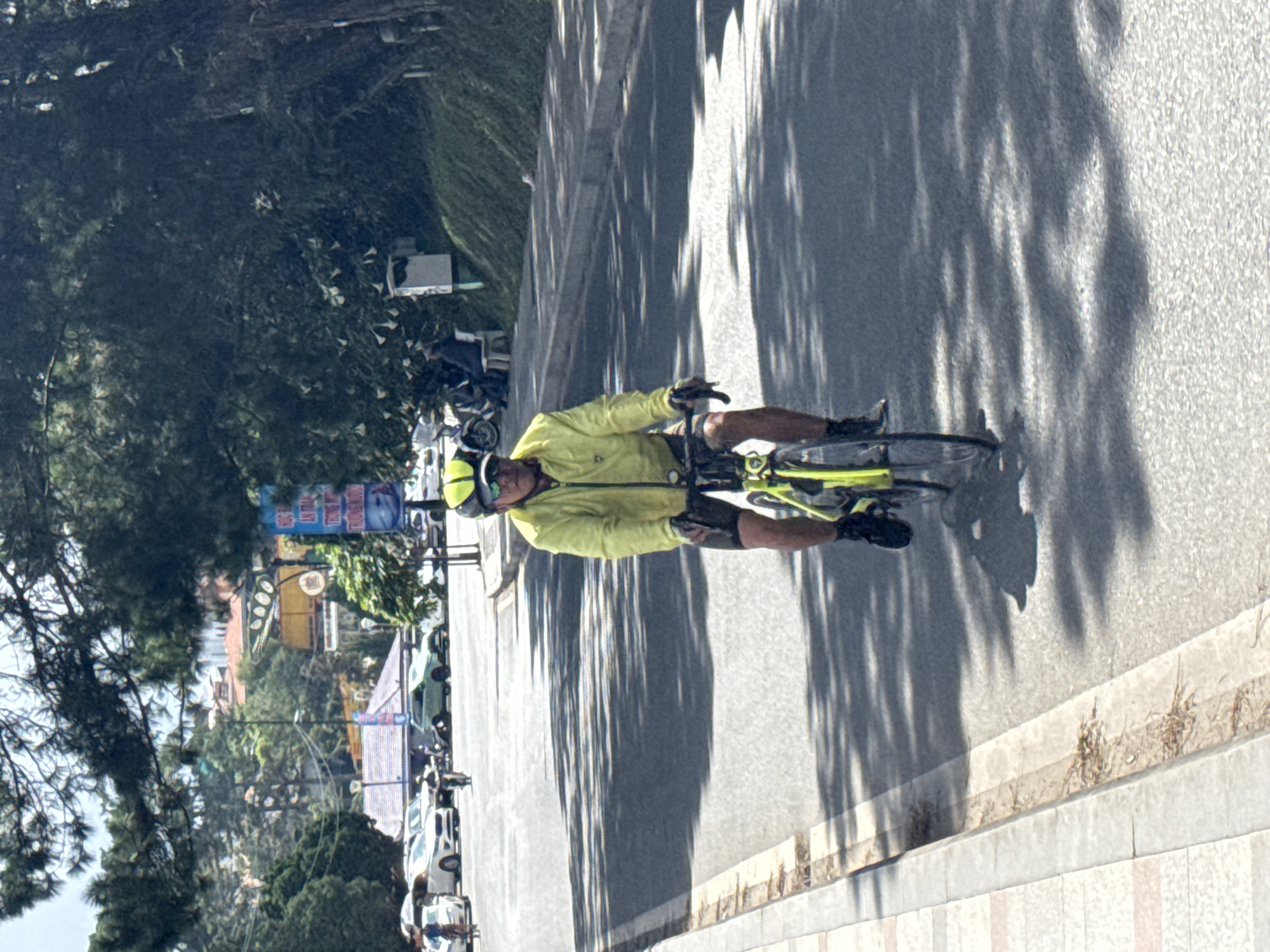 In Phường Xuân Hương - Đà Lạt, Vietnam, a man wearing a yellow cycling jacket, helmet, and sunglasses is riding a bicycle down a paved road. He is positioned in the foreground, facing forward, with the bicycle's front wheel in the lower right portion of the frame and the back wheel visible near the center. The road has a sidewalk on the left with a decorative pattern of light and dark gray and pinkish-red tiles, transitioning to a rougher gray surface near the road. Tree shadows are cast across the road, creating a dappled light effect. In the background, several cars are parked along the side of the road, including a white sedan and a light green SUV. Further back, buildings with red tiled roofs are visible among lush green trees and foliage. A blue and white sign is mounted on a pole in the mid-background. The sky is bright and appears to be daytime.