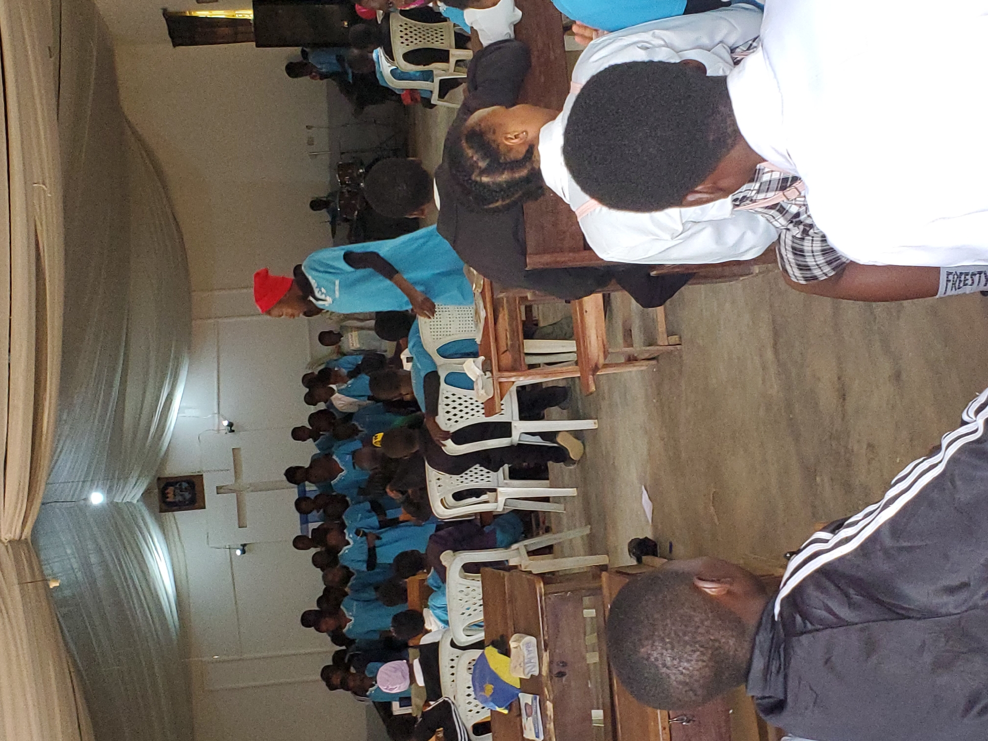 The image depicts an indoor gathering of a large group of people, predominantly young individuals, seated in rows of chairs. The scene appears to be a church or a similar place of worship due to the presence of a cross on the wall and what looks like a framed picture. Many individuals are wearing light blue shirts or uniforms, suggesting a group affiliation or event. Some are wearing head coverings, including a red headscarf on one person in the foreground and a light purple one on another.
The individuals are engaged in an activity, with most looking downwards, possibly in prayer, listening to a speaker, or participating in a lesson. The lighting suggests it could be daytime, but it's hard to determine the exact time. The overall atmosphere seems to be one of solemnity and focus.
Notable details include the mix of white plastic chairs and wooden benches, indicating a potentially makeshift or multipurpose space. The curtains on the left side of the image are thick and draped, possibly to control light or for decoration. On the right side of the image, one person is visible wearing a black jacket with white stripes on the sleeves, and another person is wearing a white top.
There is visible text on the shorts of the person in the white top, reading "FREESTY". The overall impression is of a community event or religious service taking place in Kuru Karama, Nigeria.