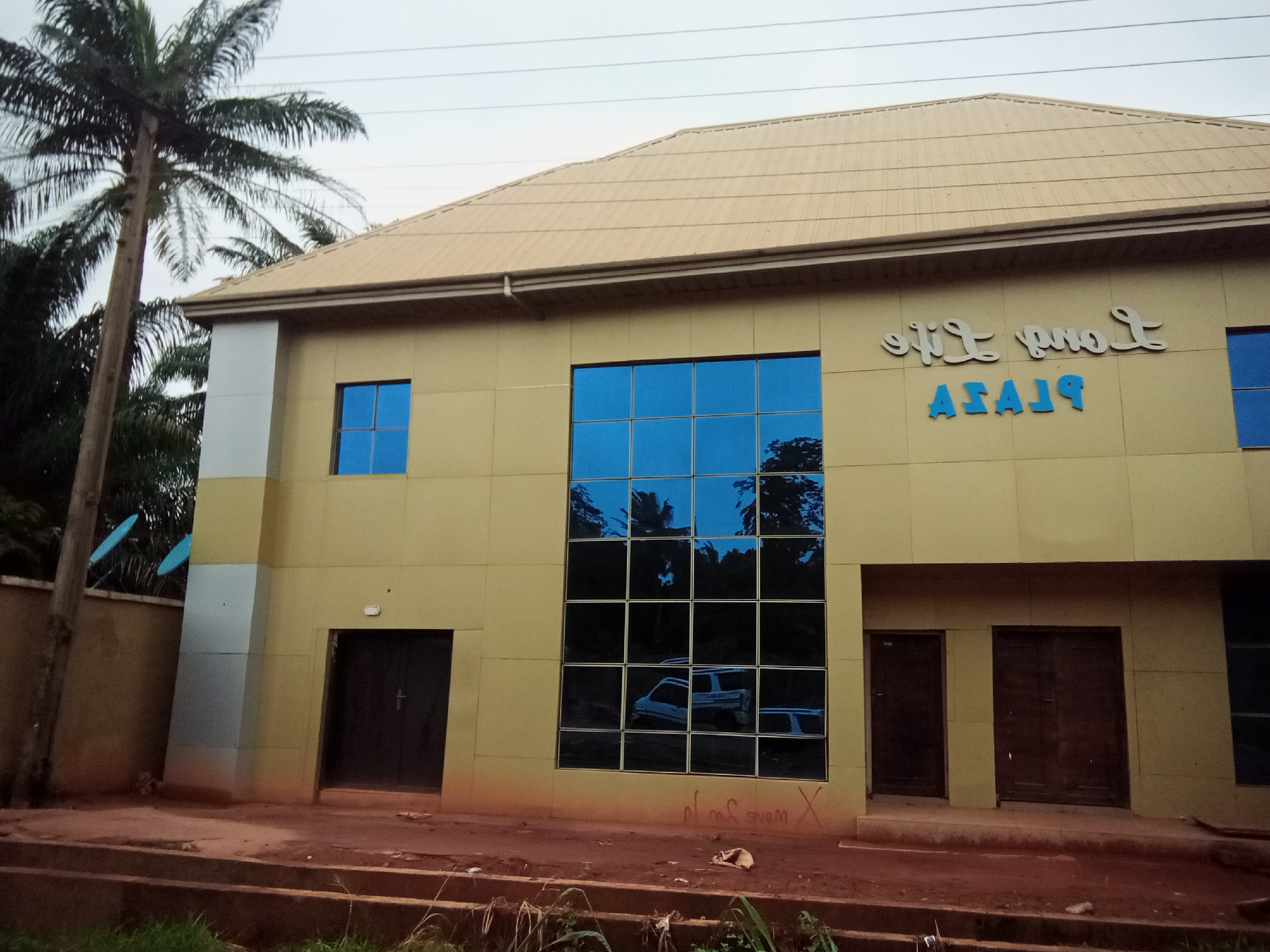 In the city of Oko Town, Nigeria, a two-story building with a beige facade stands prominently under a grey sky. The building features a tan, corrugated metal roof. On the upper facade, two signs are visible: one reads "Love Life" in cursive script, and below it, "PLAZA" is written in blue block letters. The building has several blue-tinted windows, including a large, multi-pane window on the right side of the upper floor, which reflects trees and possibly vehicles. On the ground floor, there are two dark wooden doors on the right side of the building. To the left, there is a smaller, single dark wooden door. Adjacent to the building, a tall palm tree stands to the left, its fronds extending towards the building. A beige concrete wall with two small satellite dishes is visible behind the palm tree. The ground in front of the building is a reddish-brown, possibly dirt or clay, with some grass and debris scattered around.
