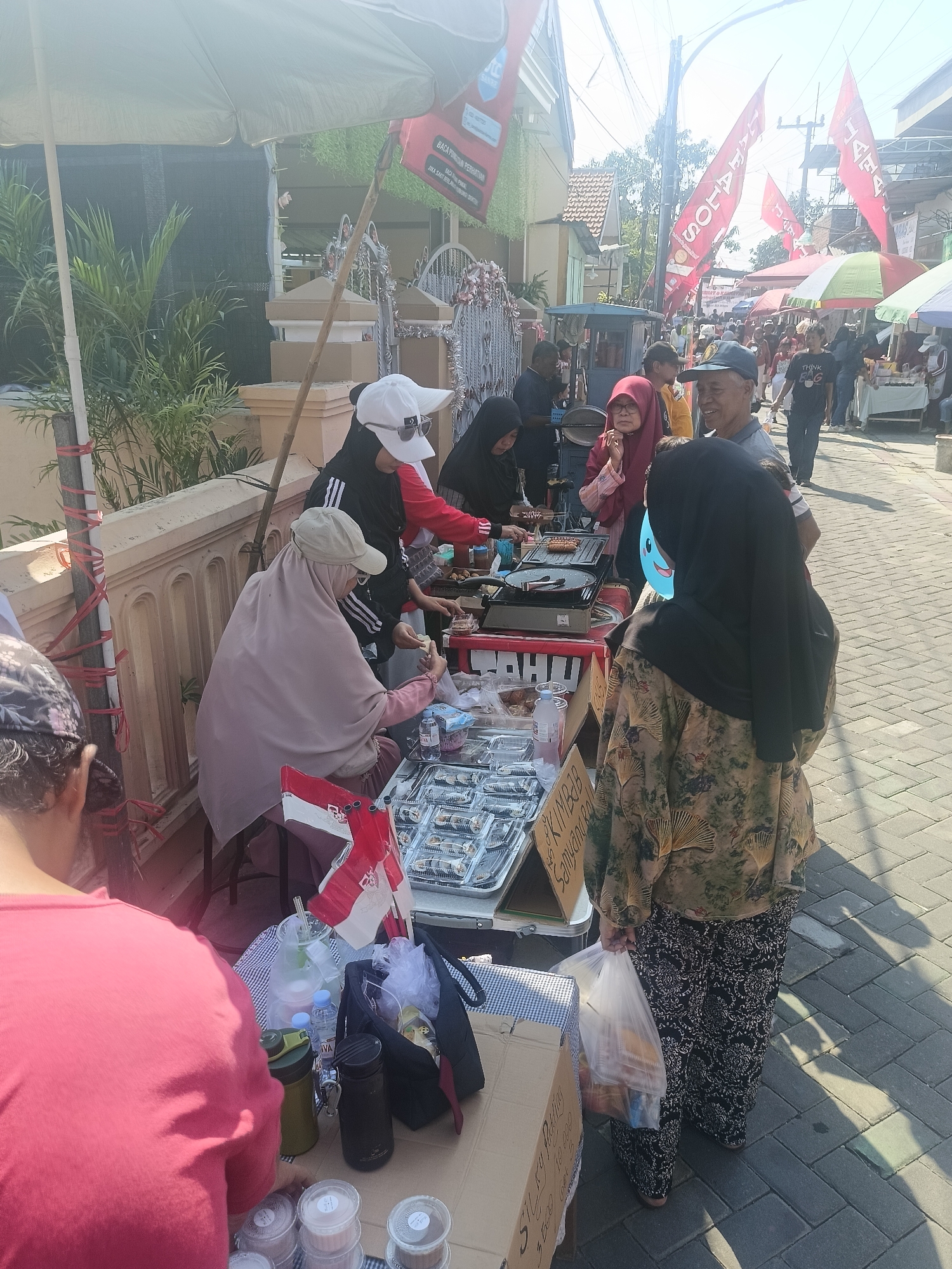 This is an eye-level, medium shot of a street scene in Gedangan, Indonesia, likely during a market or event. The foreground features a stall with various food items for sale. A woman in a pink headscarf and a beige abaya is seated behind the stall, attending to customers. To her right, a person in a black hijab and a patterned tunic is holding a plastic bag filled with goods. In the background, a bustling street is visible, with people walking and browsing other stalls. Red flags with Indonesian text are prominently displayed, suggesting a festive or national occasion. The overall atmosphere appears to be lively and communal.