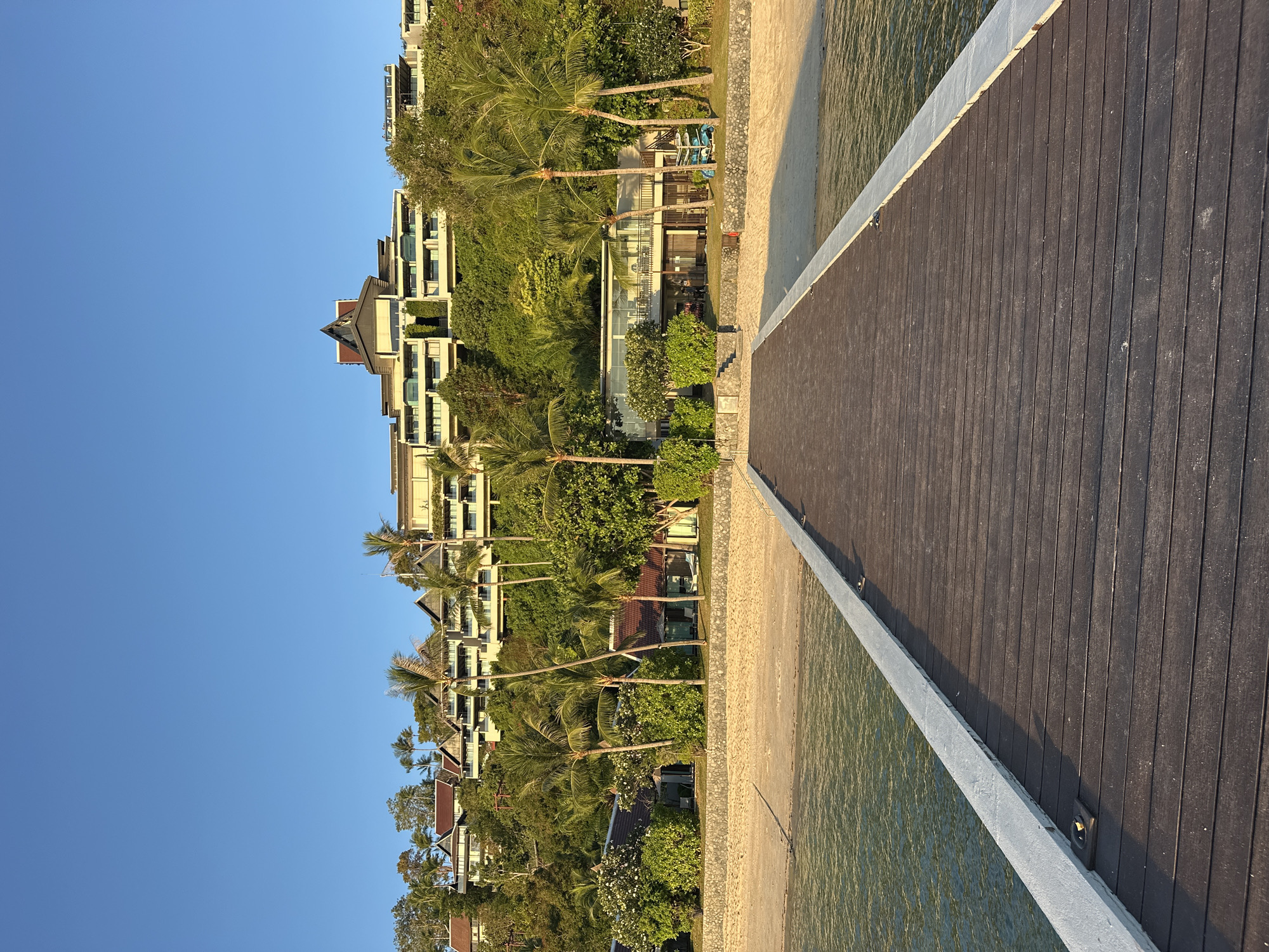 A wooden pier extends from the foreground into a body of calm, clear water with a sandy beach to the right. Beyond the beach, a resort complex is visible, built into a lush, green hillside under a clear blue sky. The resort features multiple levels of white buildings with dark roofs, interspersed with palm trees and other tropical foliage. A few smaller buildings with tiled roofs are situated closer to the beach, with some lounge chairs visible on the sand. The overall scene depicts a serene tropical resort environment.