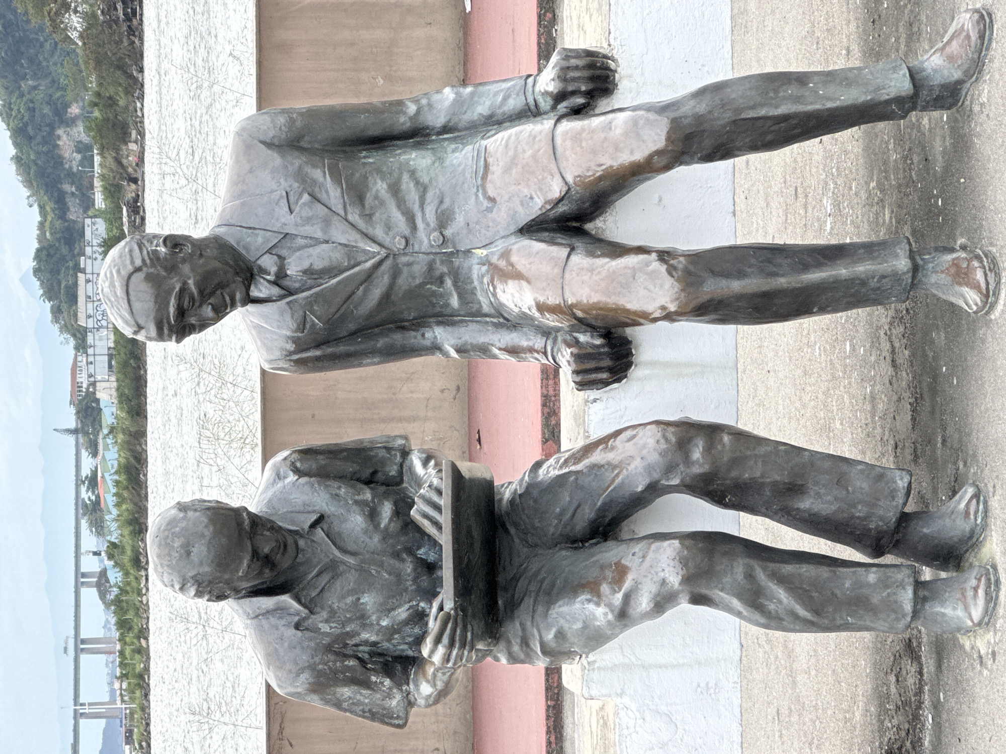 This is a full, eye-level, outdoor shot of two bronze statues of men sitting side-by-side on a low white ledge. The statue on the left is looking down and appearing to write in a ledger or book. He is wearing a short-sleeved collared shirt and pants. The statue on the right is looking towards the left statue and is wearing a suit jacket, shirt, and tie. Both statues have wet surfaces, indicating recent rain or cleaning. Behind the statues is a textured wall with a reddish-brown upper section and a lighter-colored lower section. The ground in front of the ledge is concrete. In the background, a body of water is visible, with a large bridge spanning it in the distance. Beyond the water and bridge, there are buildings and hills, characteristic of the city of Niterói, Brazil. The sky is overcast.