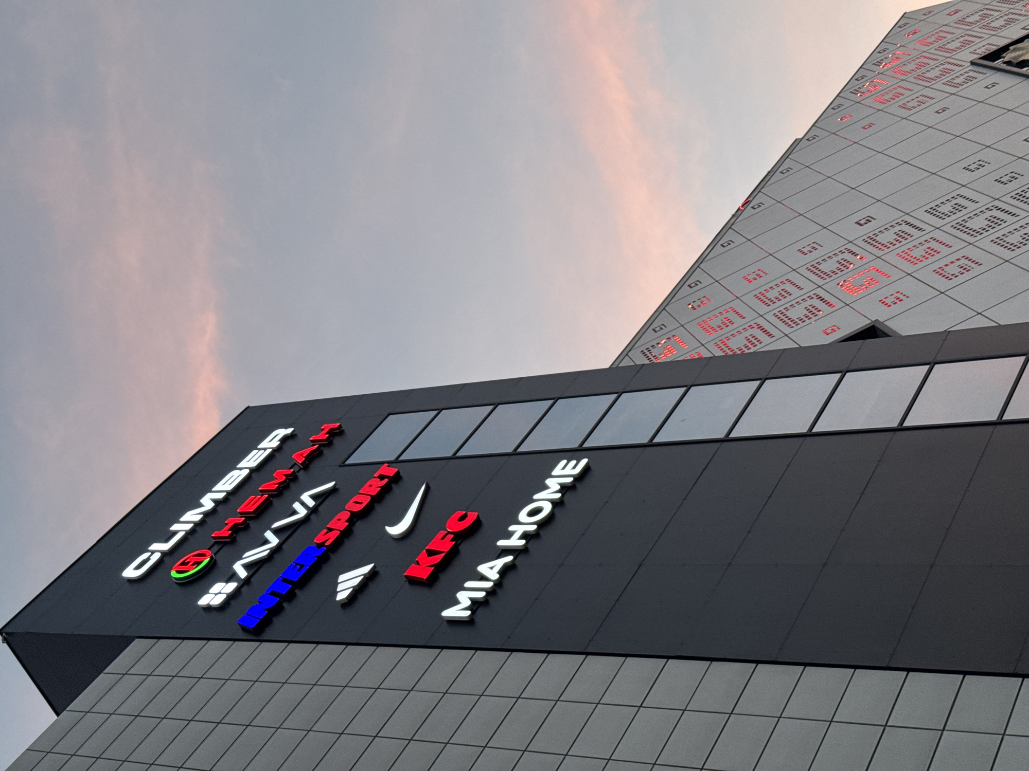 The image shows the exterior of a modern building with several illuminated signs for various businesses. On the dark facade of the building, from top to bottom, are signs for "CLIMBER," "HEMAH," "SAVVA," "INTERSPORT," "ADIDAS" (represented by its logo), "NIKE" (represented by its logo), "KFC," and "MIA HOME." To the left of the main facade, a section of the building is covered in grey rectangular panels. Another part of the building, visible on the right, is a slanted surface covered in a diamond-shaped pattern with red lights emanating from within the shapes. The sky above is a soft pink and blue, suggesting it is either sunrise or sunset. The building is identified as being located in Bishkek City, Kyrgyzstan.