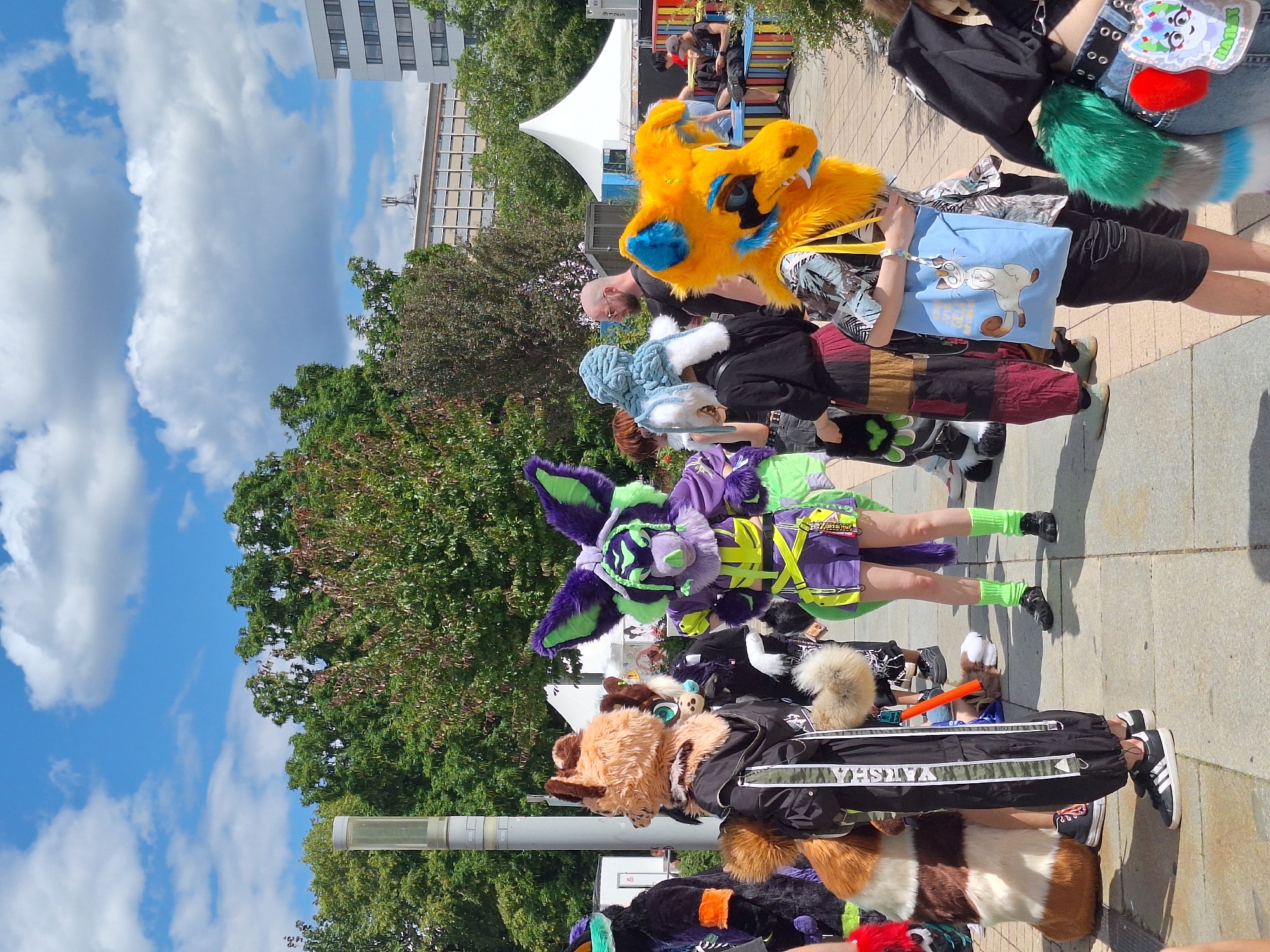The image depicts an outdoor gathering of individuals in furry costumes on a paved walkway. The setting appears to be a public space in Chemnitz, Germany, with buildings and trees visible in the background. The sky is blue with scattered white clouds.
In the foreground, several people are wearing elaborate furry costumes, characterized by animal-like heads, tails, and fur. One individual on the far left wears a black costume with orange accents and a green mask. To their right is a person in a brown and white furry costume with a long tail, wearing black pants and sneakers. Another person in a purple and green costume with large ears and a neon yellow harness is prominent in the center. To the right of this figure is a person in a blue and white fluffy headpiece. Further to the right, a person wearing a bright orange and blue furry headpiece is visible, carrying a light blue tote bag with a cartoon cat design. A person in a black outfit with green and grey furry accessories is on the far right.
Other individuals, some without costumes, are present in the middle ground and background. Some are seated on a colorful bench structure in the background, while others are standing. A white tent is set up behind the costumed individuals, suggesting an event or convention. Trees line the background, and a tall, modern building is visible on the right side of the image. The overall scene suggests a community event or gathering focused on furry culture.
