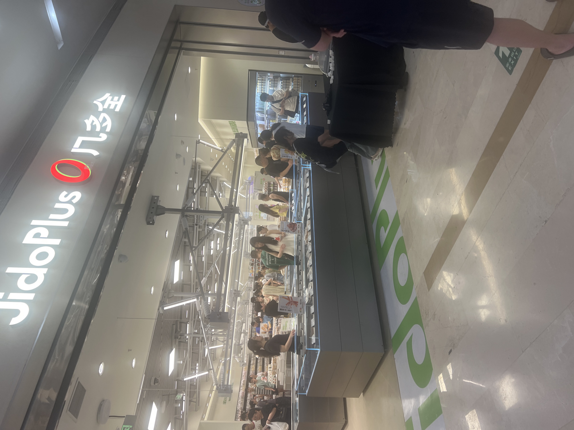 This is a medium shot of the interior of a brightly lit, modern supermarket. The store features a large, illuminated sign above the entrance that reads "JidoPlus 凡多全". Inside, various display cases are visible, stocked with items, and several shoppers are browsing. The floor is polished marble with a green directional arrow graphic on it. In the foreground, a black fabric-covered table is set up, with a few white cube-shaped objects on top. A person in a dark t-shirt and shorts is standing near the table, partially obscuring it. The overall impression is a busy retail environment. This image was taken in Liandu District, China.