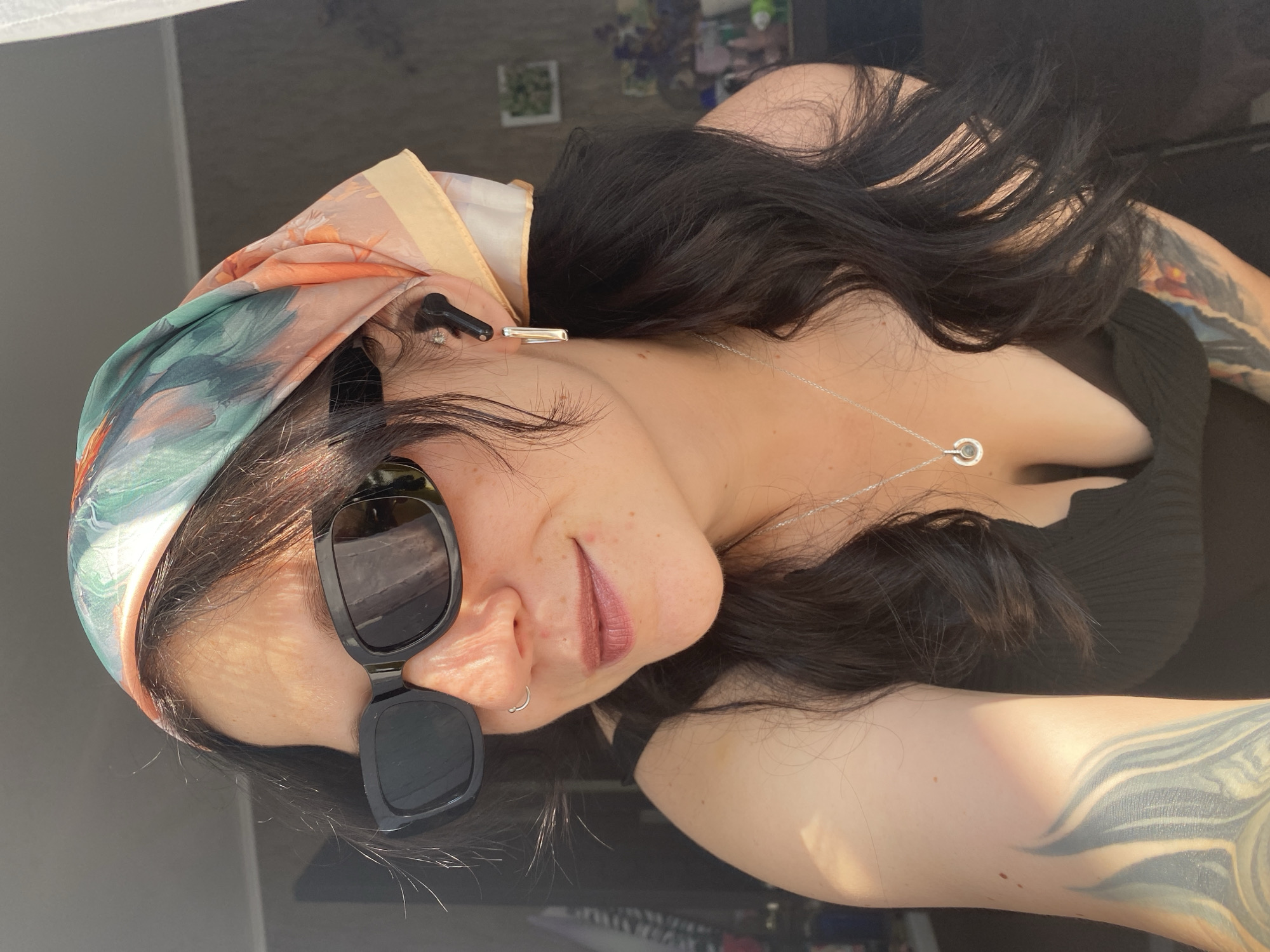 The image is a close-up, outdoor, selfie-style portrait of a woman wearing a headscarf and sunglasses. She has dark hair, visible tattoos on her arm and chest, and is wearing a black top, a silver necklace with a circular pendant, and hoop earrings. She also has a nose ring. She is wearing black wireless earbuds. The lighting suggests it is daytime, with sunlight casting shadows and highlighting her face. The background is blurred and appears to be an indoor setting with some plants and objects visible.
There is no information in the image to indicate its location in the city of Chaikovsky, Russia.