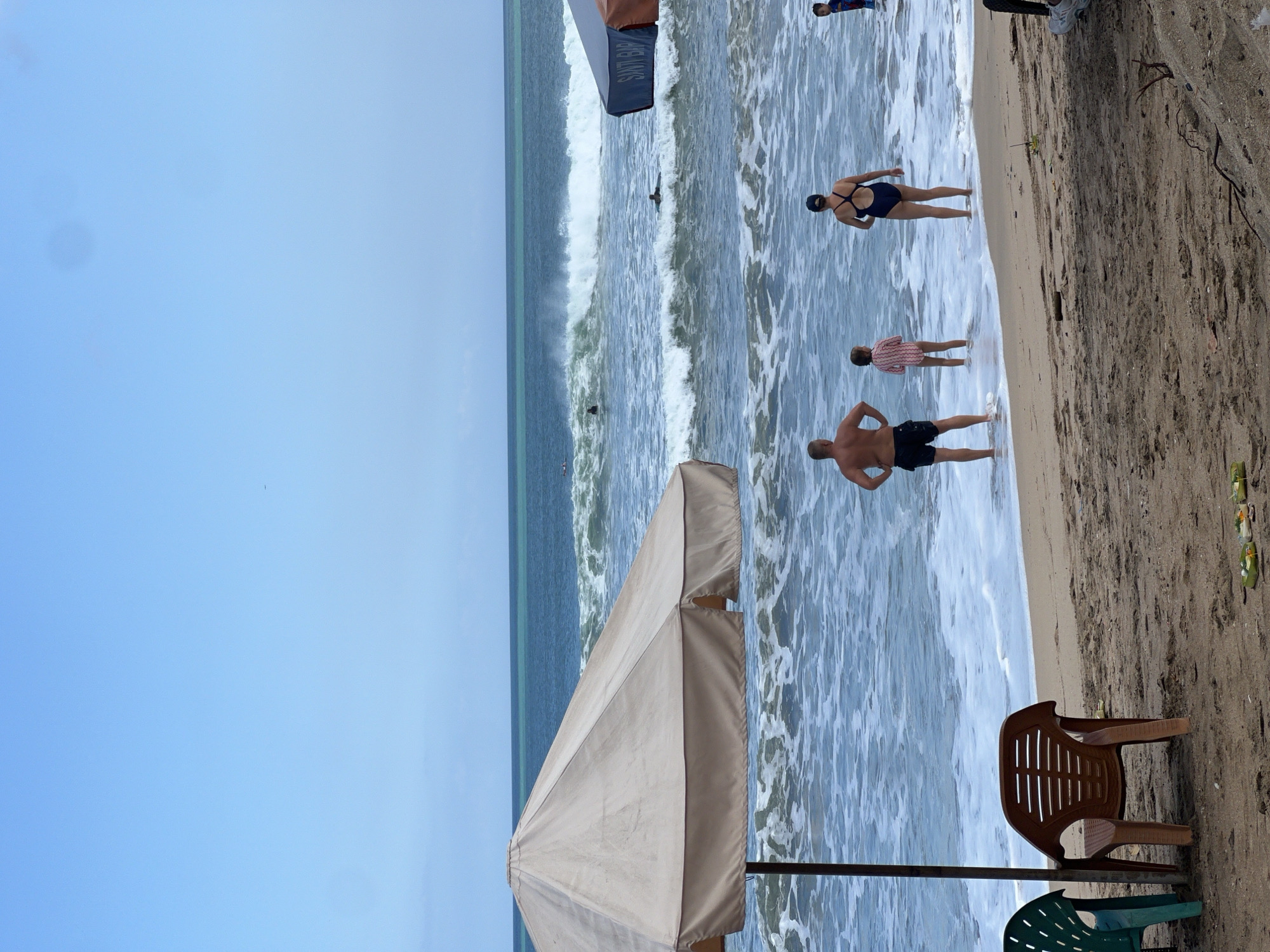 On a sandy beach in Kuta, Indonesia, several people are interacting with the ocean. In the foreground, a large tan beach umbrella shades a portion of the sand. Two empty plastic chairs, one green and one brown, are positioned on the sand near the umbrella. Further down the beach, a man wearing black swim trunks stands facing the ocean. Beside him, a young girl in a striped swimsuit stands in the shallow water. To the right of the girl, a woman in a navy blue swimsuit is in the water. In the background, the ocean is active with moderate waves. Several surfers are visible in the water, some riding waves and others paddling. A second, smaller beach umbrella with the words "SANTI BAR" visible is partially in the frame on the right. The sky is clear blue with a few faint clouds.