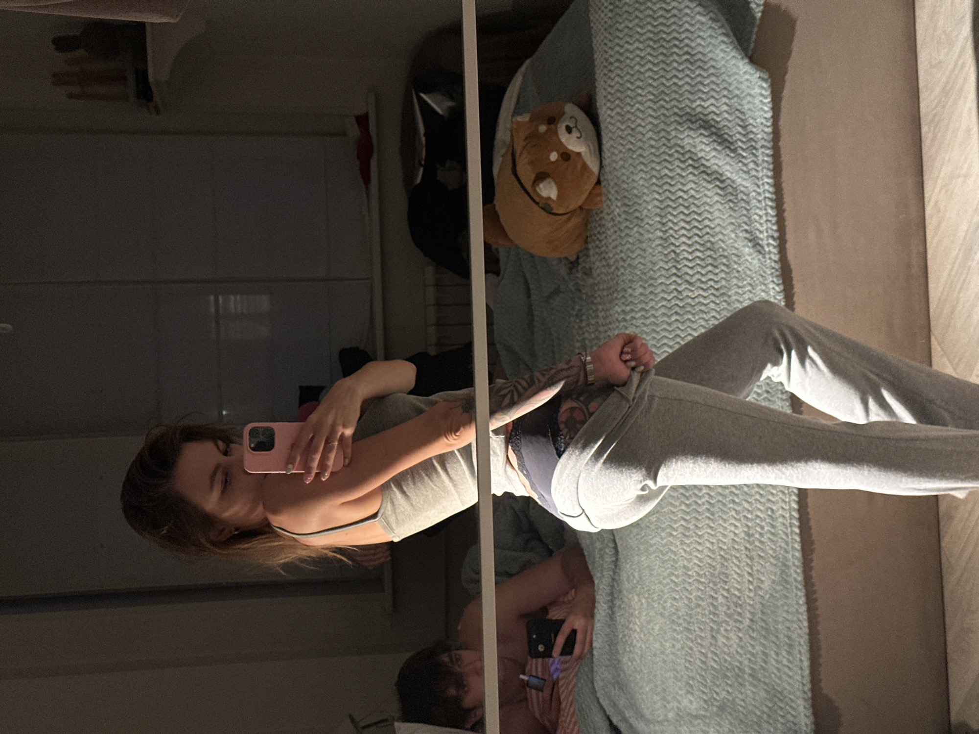 In a dimly lit bedroom, a young woman with light brown hair, wearing a grey tank top and grey sweatpants, is taking a selfie in a mirror. She holds a pink phone in her right hand, and her left hand is pulling at the waistband of her sweatpants, revealing black lace underwear. She has visible tattoos on her left arm. In the background, a young man lies on a bed, looking at his phone, with a cigarette in his right hand. A light blue textured blanket covers the bed, and a stuffed animal resembling a Shiba Inu dog is placed on the bed. A radiator is visible behind the bed, and a shelf with objects is on the wall to the right. The overall scene suggests a casual, indoor setting, possibly a bedroom in a residential apartment.