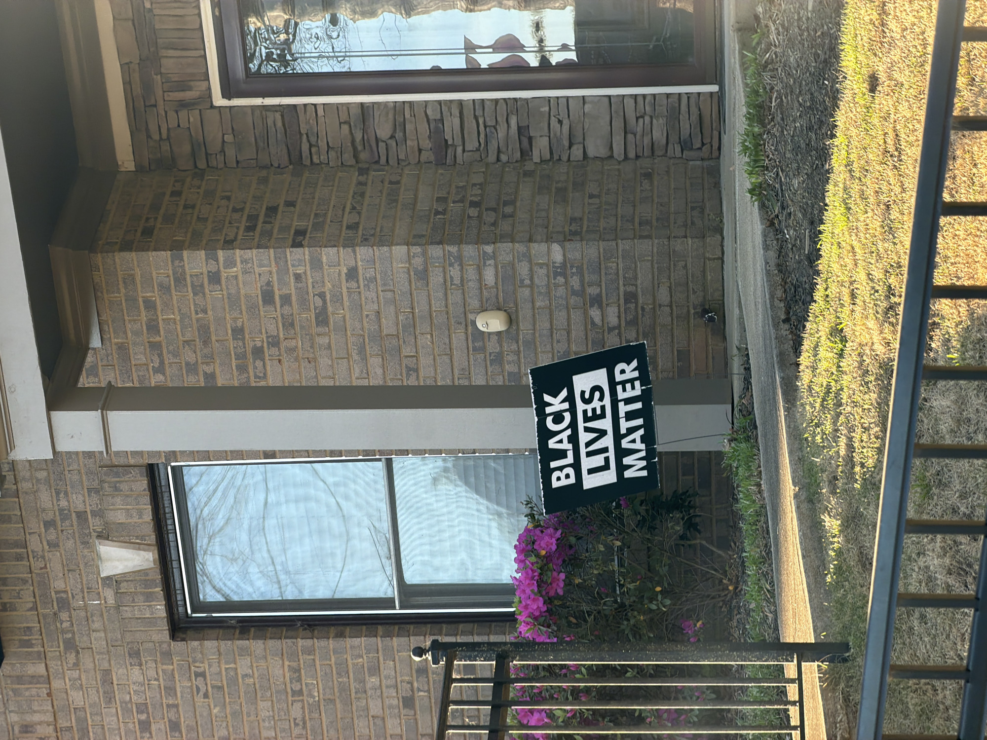 A "Black Lives Matter" sign stands near a brick-walled house in an outdoor setting. The house features windows with visible reflections and a light fixture on the brick wall. A small bush with pink flowers grows near the sign. A metal fence is partially visible in the foreground. Grass and greenery are present on the right side of the frame. The location is presumed to be in a residential area within the city of Atlanta, United States of America, based on visual cues such as architectural style and landscape. There are no people present in the image.
