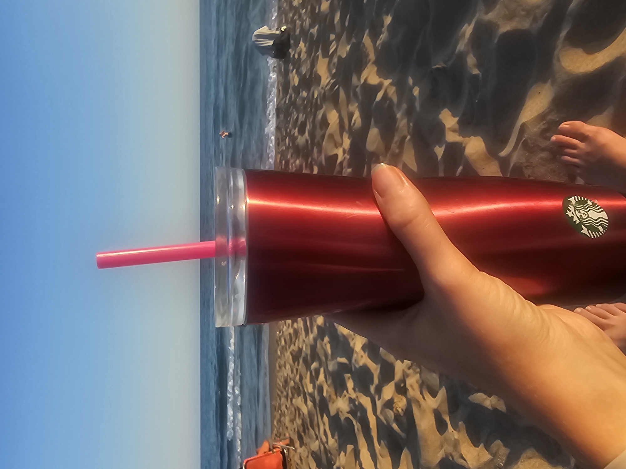 This is a vertically oriented, eye-level, first-person perspective medium shot of a person holding a red reusable tumbler with a pink straw on a sandy beach. The tumbler has a Starbucks logo on it. The person's hand is visible holding the tumbler, and their feet are also visible in the foreground. The background consists of the ocean and a clear blue sky, suggesting a sunny day. The lighting indicates it is likely late afternoon or early evening, given the long shadows cast on the sand. The scene appears to be a relaxing vacation or outing at the beach.