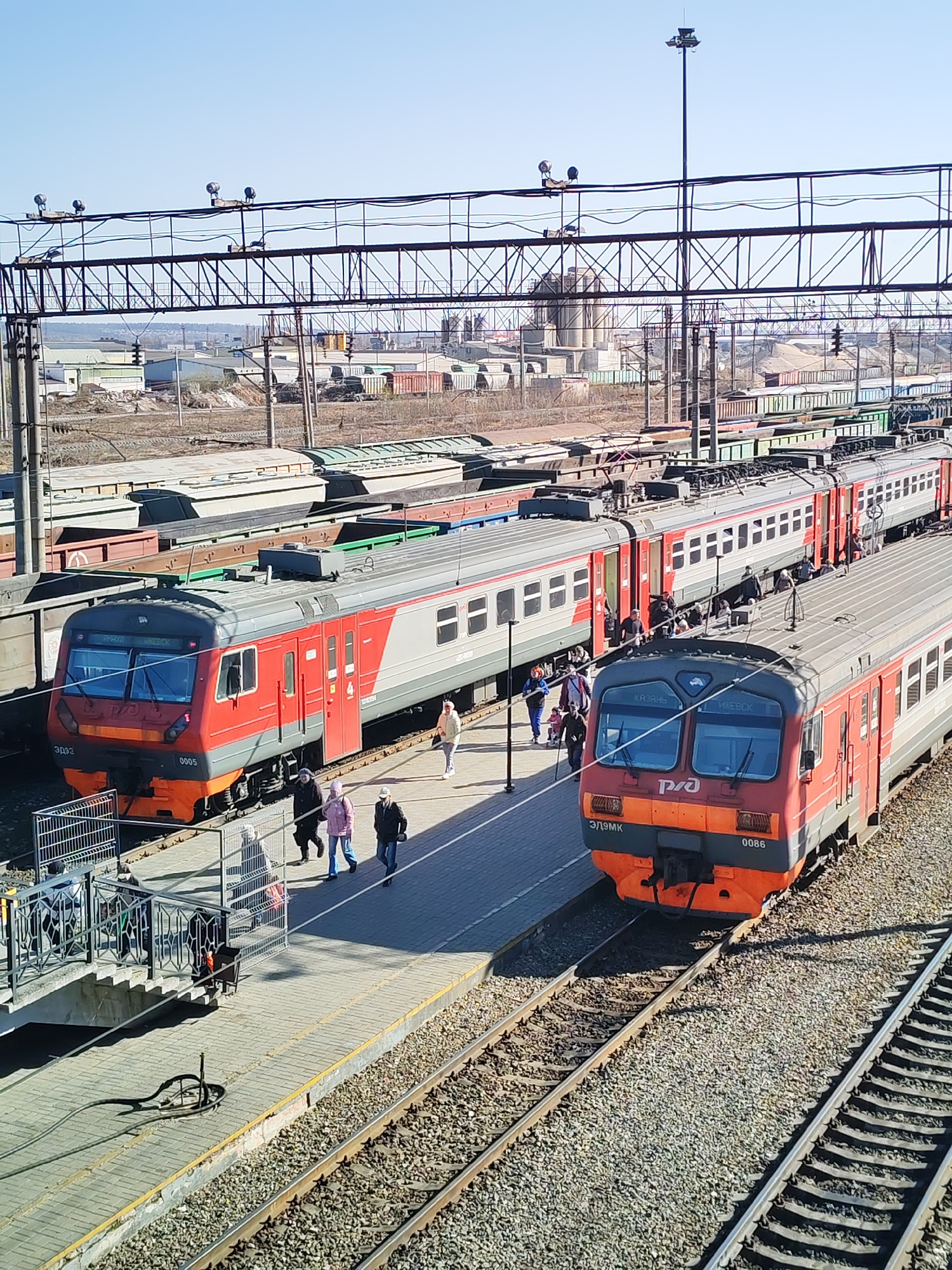 The image displays a bustling railway station or yard in Izhevsk, Russia, under a clear, sunny sky, suggesting daytime. The scene is dominated by two red and grey electric multiple unit (EMU) passenger trains.
The train positioned at the platform, to the left, is an "ЭД9Э" model, numbered "0005", and prominently features the Russian Railways (РЖД) logo. Its destination display shows "УРЖУМ" and "ИЖЕВСК". On a parallel track to the right, another similar train, an "ЭД9МК" model, numbered "0086", also carries the РЖД logo and displays "КАЗАНЬ" and "ИЖЕВСК" on its front.
Multiple people are visible on the platform, engaged in activities typical of a train station, such as walking and standing, likely boarding, disembarking, or waiting for trains. Their attire, including light jackets and hoodies, suggests cool or mild weather. A pedestrian overpass with stairs provides access to the platform.
In the background and mid-ground, numerous freight wagons, both covered and open-top, are parked on various tracks, indicating a large rail hub handling both passenger and cargo transport. Overhead gantries with lights and catenary structures span across the tracks. Industrial buildings, silos, and what appear to be storage containers are visible in the distance. The tracks are laid on gravel ballast, and a coiled cable or hose lies on the platform near the overpass stairs. The overall impression is a routine, active day at a regional train station.