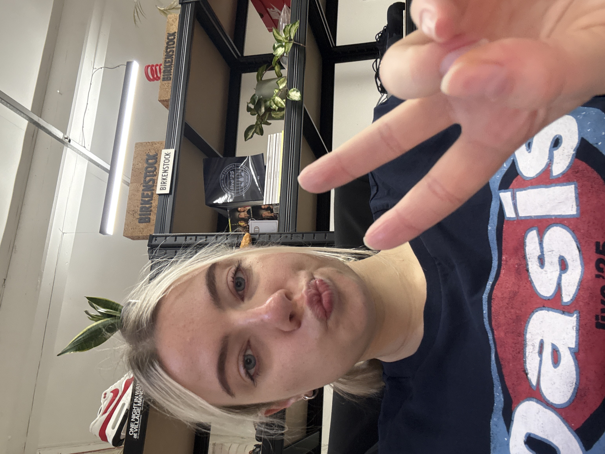 A young, blonde-haired woman is taking a selfie. She has her mouth pursed and is holding up her fingers, potentially forming a peace sign. She is wearing a dark blue t-shirt with a graphic print that reads "Oasis live '95" in white and red letters with a blue outline. Behind her, a shelf unit is visible, holding various items including boxes with "Birkenstock" branding, books, and a potted plant. There's also a rectangular light fixture on the left side of the image.
Based on the "Oasis live '95" t-shirt, the location is potentially somewhere connected with the band Oasis, who are famously from Manchester, United Kingdom. Without further details, it is impossible to pinpoint the exact location.
