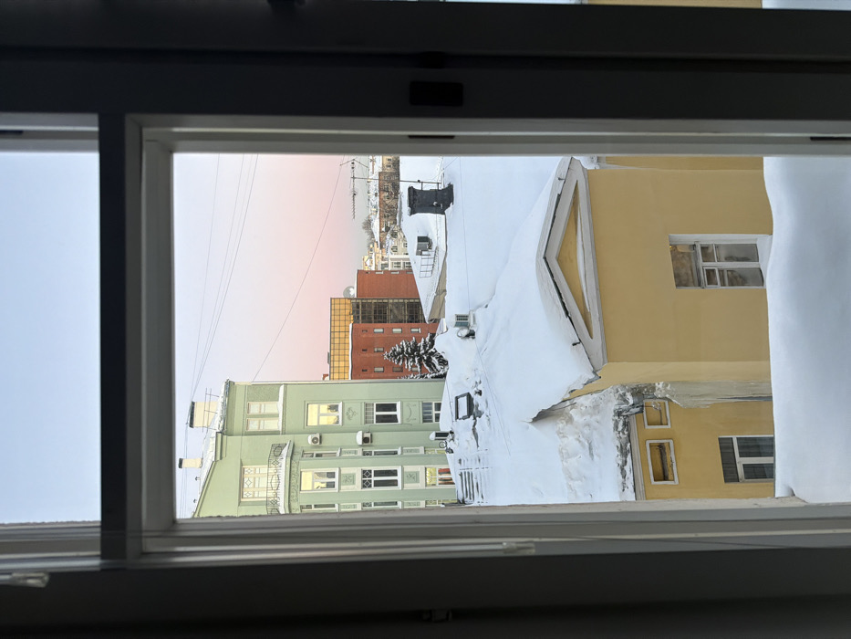 The image depicts a winter cityscape viewed through a white window frame, offering an elevated perspective of a residential and urban area in Moscow, Russia. The scene is characterized by extensive snow cover on all visible rooftops and ground surfaces.
In the midground, a prominent yellow two-story building features a classical pediment-like facade element on its roof and multiple rectangular windows. Its roof is heavily blanketed in snow, and large icicles hang from the eaves on the left side. Behind and to the left of this yellow structure stands a multi-story, light green building, identified as located at Prechistenka Street 20. This building exhibits intricate architectural details, including a rounded corner with a small balcony and numerous windows, some of which reflect the light. Several outdoor air conditioning units are affixed to its lower facades. Its roof is also snow-covered. Adjacent to the green building, a snow-laden dark green fir tree is visible.
In the background, a mix of urban architecture includes red and orange multi-story buildings, some featuring modern glass facades. Distant rooftops, also covered in snow, stretch towards the horizon. Electrical wires crisscross the sky, and a television antenna array is visible on a distant rooftop. The sky above is clear, displaying a gradient from light blue at the zenith to a soft pale pink and orange hue near the horizon, suggesting either dawn or dusk. No individuals or active interactions are visible in the scene.