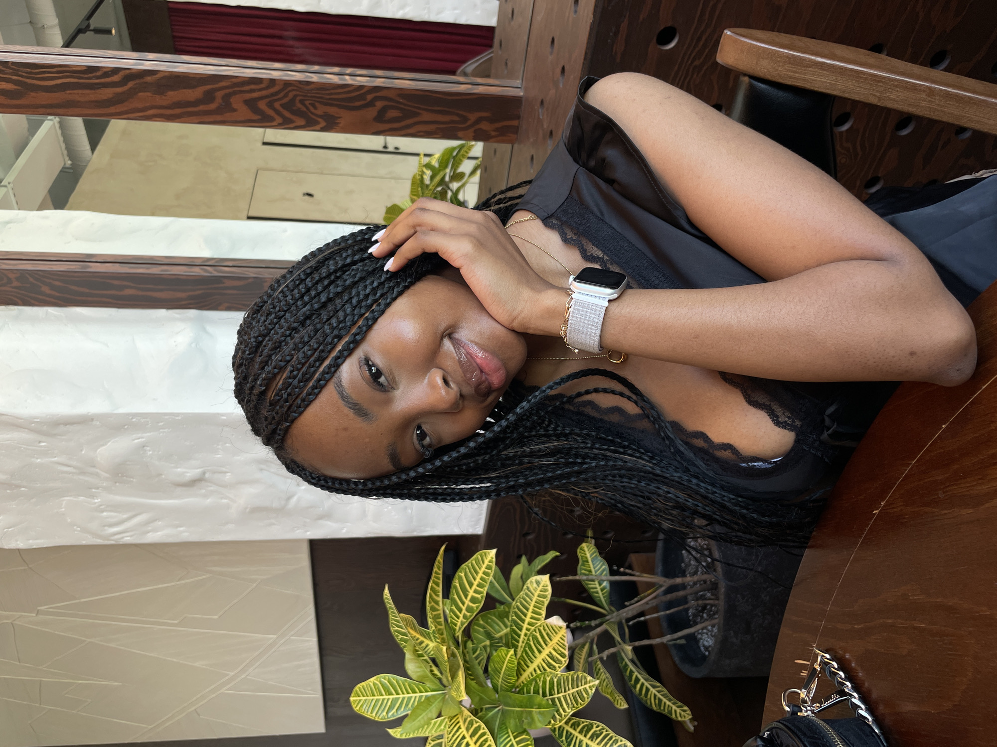 A young Black woman with braided hair is seated at a wooden table in what appears to be an indoor dining establishment. She is wearing a black top with lace trim and an Apple Watch on her left wrist. Her right hand is propped on her chin, and she is looking at the camera with a slight smile. In the background, there is a potted plant, a textured wall, and a mirror reflecting a curtain and part of the room. The lighting is natural and diffused.