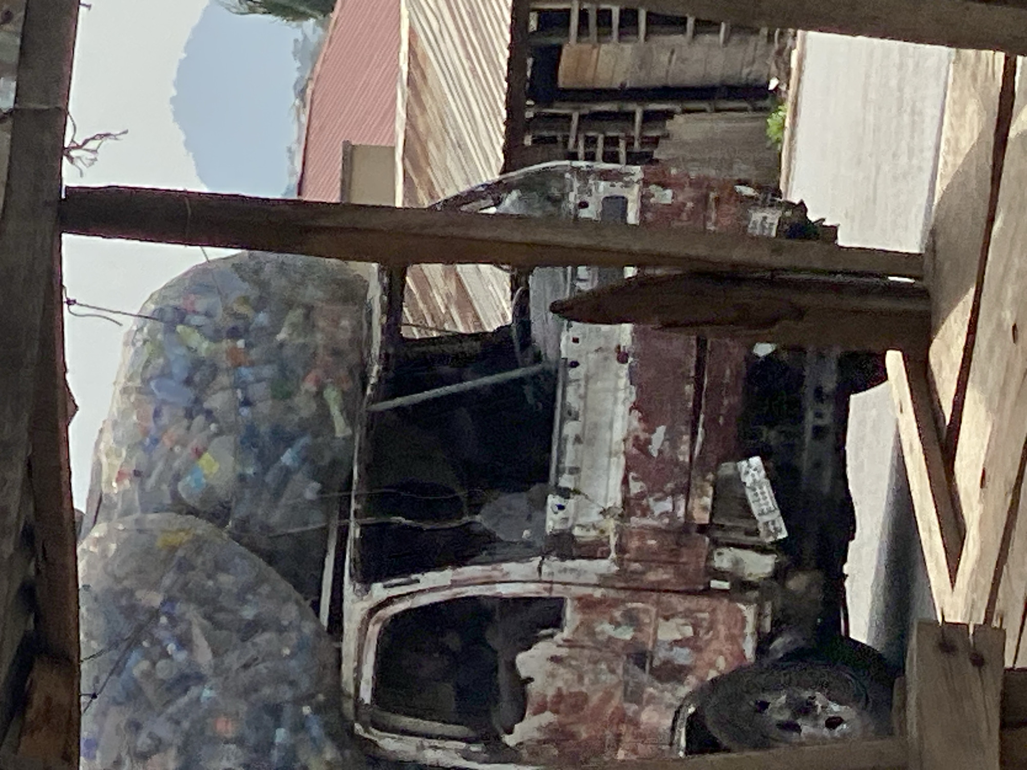 The image shows the remnants of a rusty, derelict truck in Adankolo, Nigeria. The truck's cab is heavily damaged with extensive rust and missing panels. A large net-like sack overflowing with plastic bottles is attached to the back. The scene is framed by wooden structures, suggesting it might be viewed from within a building or enclosure. The sky is visible in the upper portion. Other partially-visible buildings, possibly with corrugated roofing, are in the background.