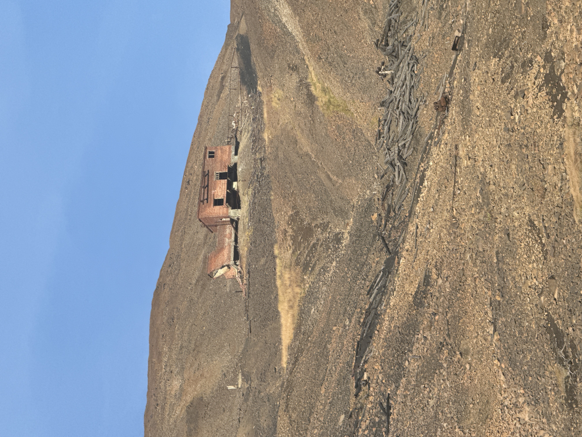 The image shows a derelict brick building situated on a barren, rocky hillside under a clear blue sky. The building, appearing partially collapsed and in a state of disrepair, has visible windows and a roof structure. The surrounding landscape is characterized by dry, uneven terrain with scattered rocks and sparse vegetation. In the foreground, a pile of weathered wooden debris lies on the ground, and further down the slope, remnants of rusty metal machinery are visible, suggesting a former industrial or mining site. The overall scene conveys a sense of abandonment and decay in a remote, rugged environment. The location is identified as Unknown, Russia.