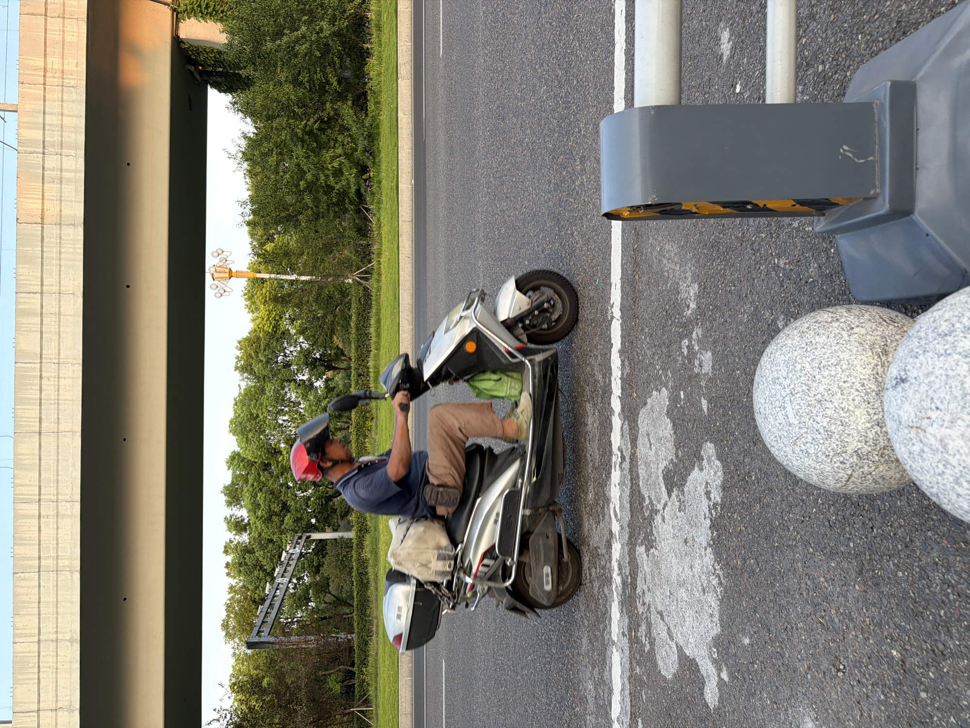 The image shows a man riding a silver electric scooter on a paved road. He is wearing a blue short-sleeved shirt, brown pants, a red helmet with a clear visor, and green shoes. He has a beige bag slung over his shoulder and a small brown pouch attached to his waist. The scooter has a black storage box on the back, and a green bag is hanging on the side. The road has white lane markings. In the background, there are trees, a street lamp with multiple bulbs, and a concrete overpass. The scene appears to be in Jiangjin, China. The lighting suggests it is daytime, possibly late afternoon, given the warm glow on the overpass. In the foreground, there are two large, round, speckled gray bollards and a gray traffic barrier with yellow and black reflective tape.