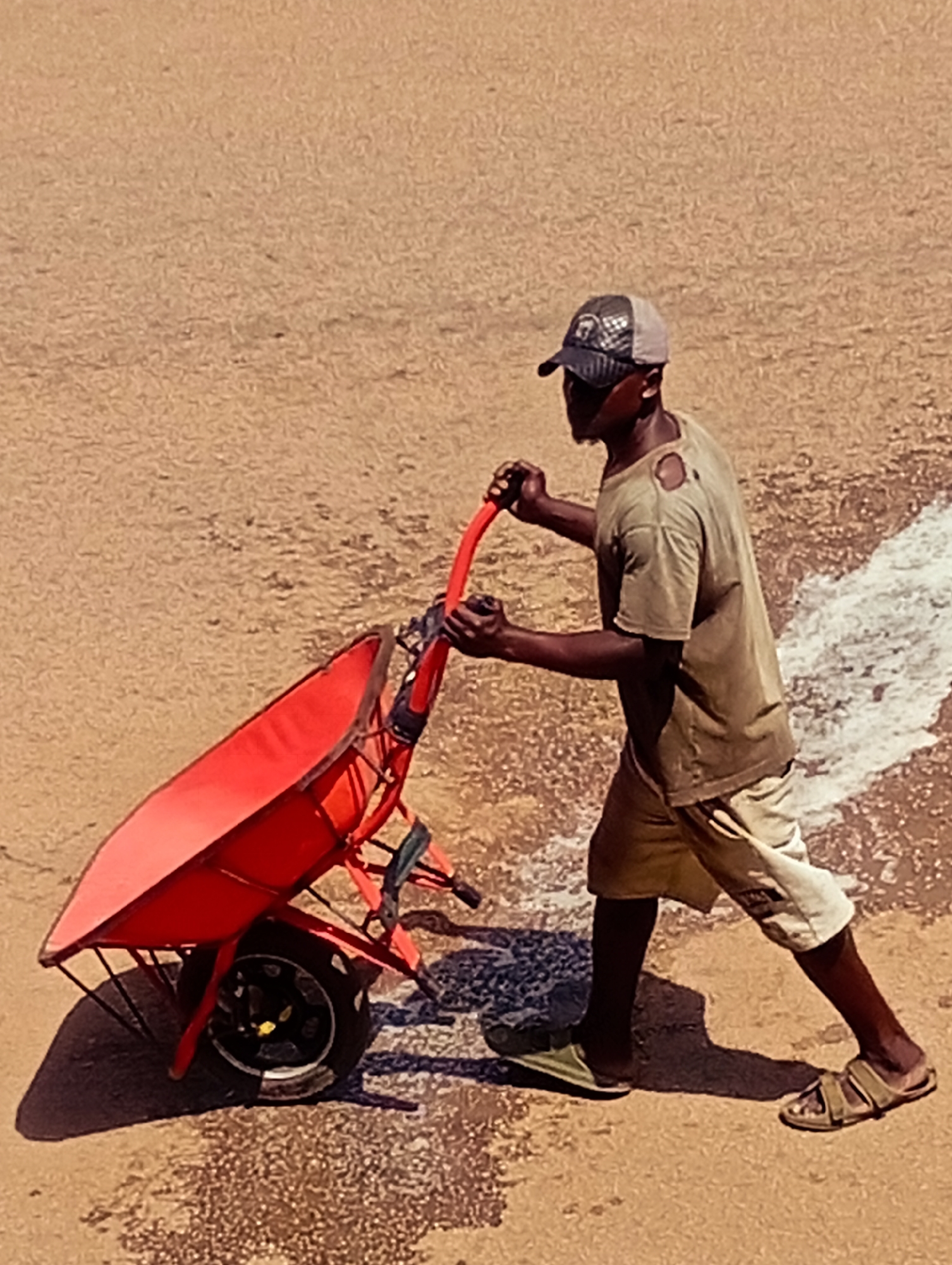 In Bukuru, Nigeria, a man wearing a cap and a torn olive green t-shirt, along with light-colored shorts and sandals, pushes a bright red wheelbarrow across a dusty, sandy surface. A white line is visible to the right of the man, and the ground appears to be rough and uneven. The wheelbarrow is tilted forward, with its single wheel in the foreground. The man's posture suggests he is in motion, likely engaged in labor or transport. The lighting casts a noticeable shadow from the wheelbarrow.