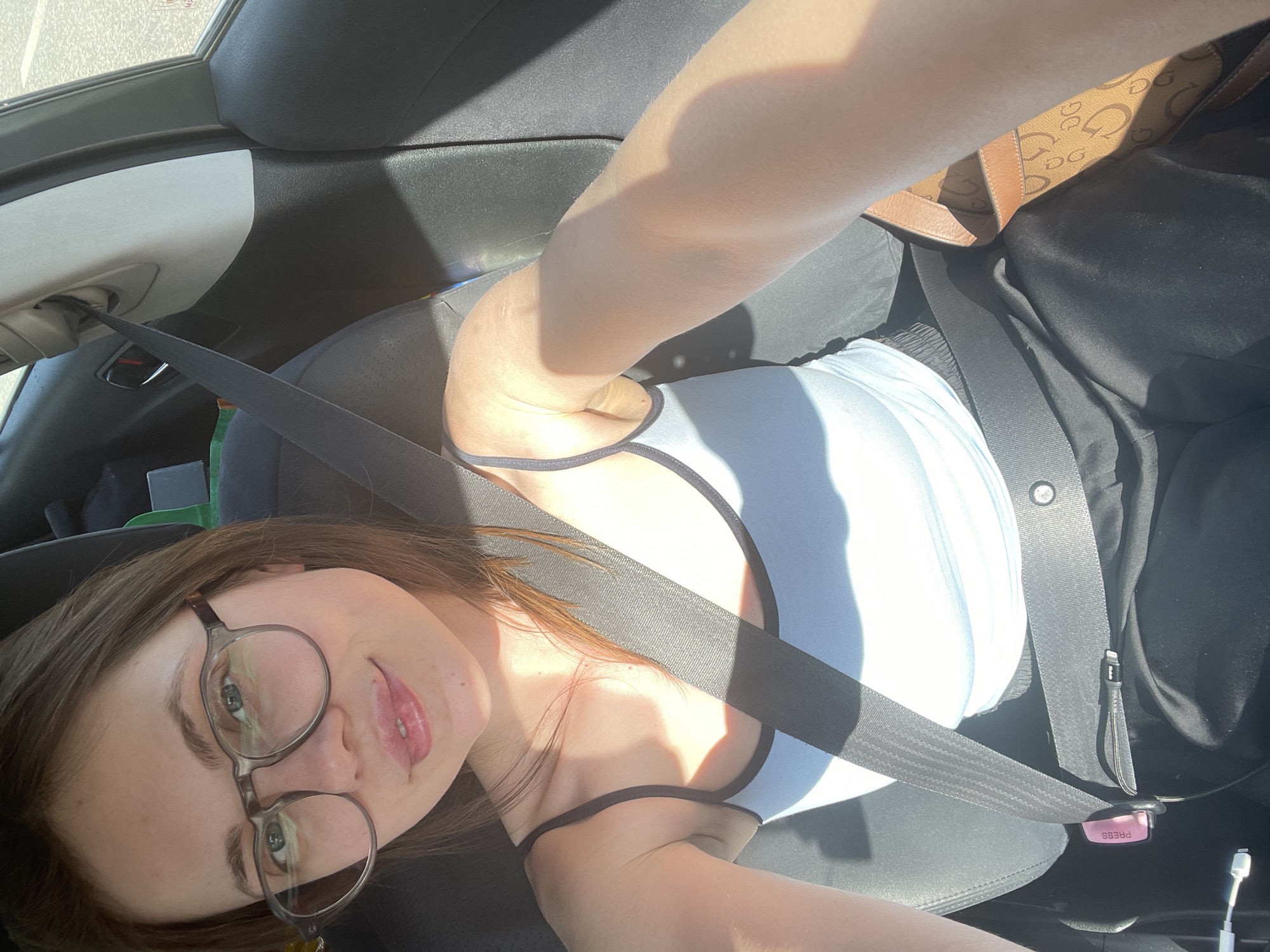 A person is in the driver's seat of a car, wearing a seatbelt. The person has brown hair, glasses, and is wearing a light blue top and black pants. A brown purse is visible in the upper right corner. The person is holding a phone and taking a selfie. The location is not identifiable from the image.
