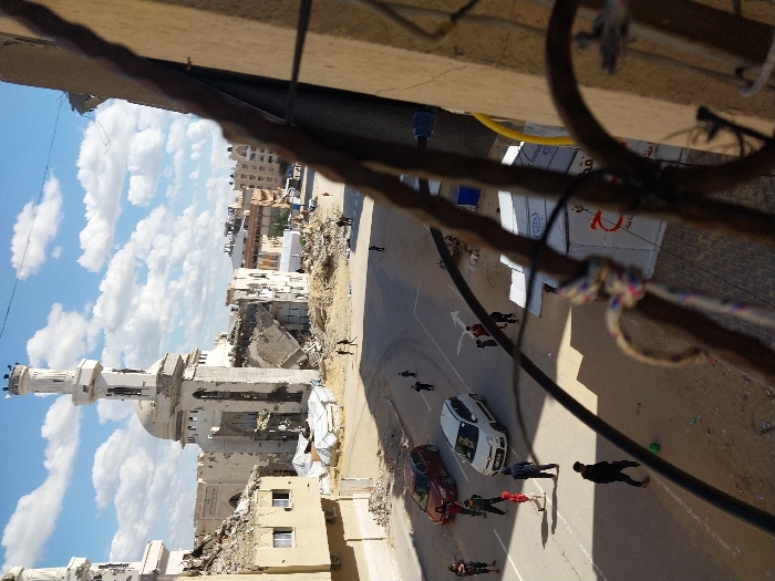 An elevated view captures a street scene in Khan Yunis, Palestinian Territory, characterized by extensive structural damage under a clear blue sky with scattered white clouds. In the midground, a prominent mosque features a tall, largely intact minaret with visible surface damage and elements on its upper platform. Adjacent sections of the mosque complex and surrounding buildings are heavily damaged, with large portions collapsed into piles of rubble and exposed interiors.
The asphalt street, marked with white arrows for traffic direction, features a white hatchback and a red SUV. Numerous individuals, identified as men and children, are present on the street and adjacent dirt areas. In the foreground, a child in a black shirt kicks a green ball, while other individuals walk or stand. To the immediate right, partially obscured by foreground wiring and a concrete pillar, are white temporary shelters displaying red Arabic text. The scene is illuminated by direct sunlight, casting clear shadows. Foreground elements consist of dark cables, wires, and a concrete building facade, indicating the photo was taken from a building.