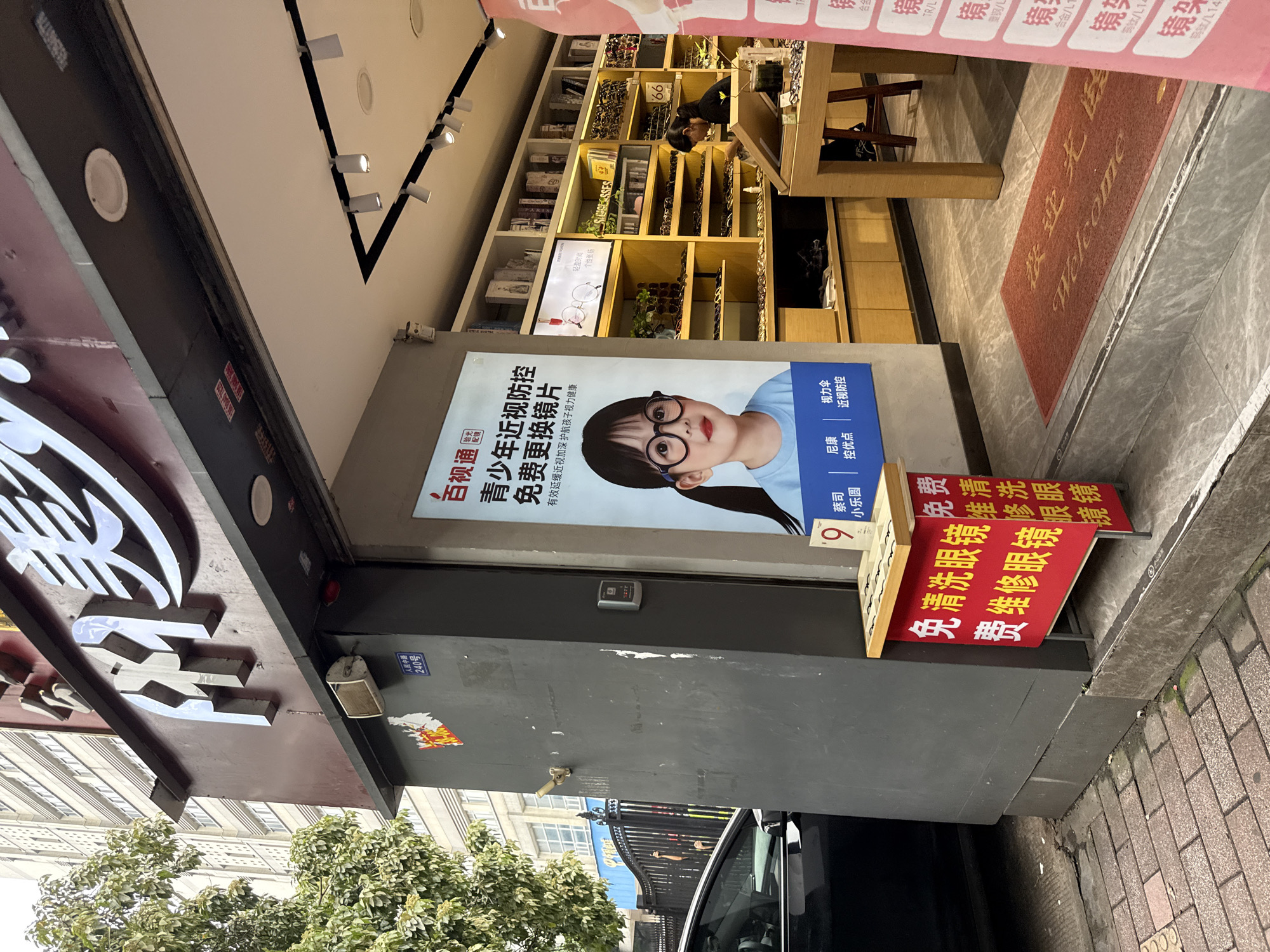 In Yuexiu District, China, a storefront for an optical shop is visible. The shop features large, illuminated signage with Chinese characters. A prominent advertisement on the left side of the entrance showcases a young girl wearing glasses, promoting a campaign for children's myopia prevention and free eyeglasses replacement. Below this advertisement, a red stand displays smaller signs offering services such as free eyeglass cleaning and repair. The interior of the shop, seen through the open entrance, reveals shelves stocked with various eyeglasses. A woman is observed inside, seemingly attending to a customer or organizing the merchandise. A red welcome mat with Chinese characters and the English word "Welcome" is placed on the floor at the entrance. The street outside the shop is paved with bricks, and a black car is parked to the left. The building to the left of the optical shop is a multi-story structure with numerous windows.