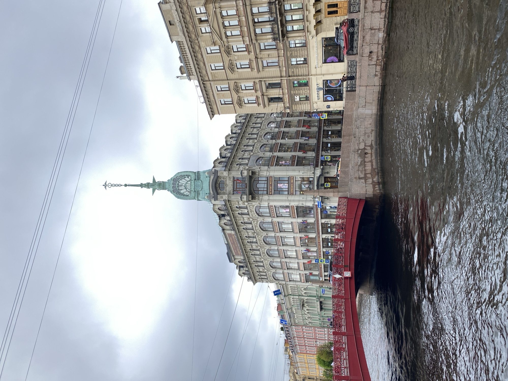 The image depicts a waterway in an urban environment with buildings lining the edge. The waterway, occupying the right side of the image, reflects the cloudy sky. On the left, buildings of various heights and architectural styles stand along the waterway. A red bridge spans the waterway. The sky is overcast. The location is Singer House, Saint Petersburg.