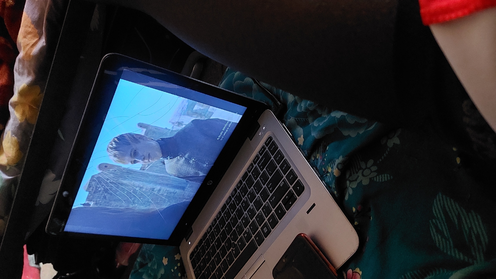 The image shows a laptop with a cracked screen displaying a scene from a show, likely Game of Thrones, featuring a young blonde character. The laptop is open and placed on a patterned fabric, possibly a blanket or bedspread, with floral designs in shades of teal and yellow. A red smartphone is visible on the left side of the laptop. The background appears to be a room, with a dark object, possibly a bag or furniture, on the top right. The lighting suggests an indoor environment. The image's context of New Nyanya, Nigeria, is not directly evident from the visual elements.