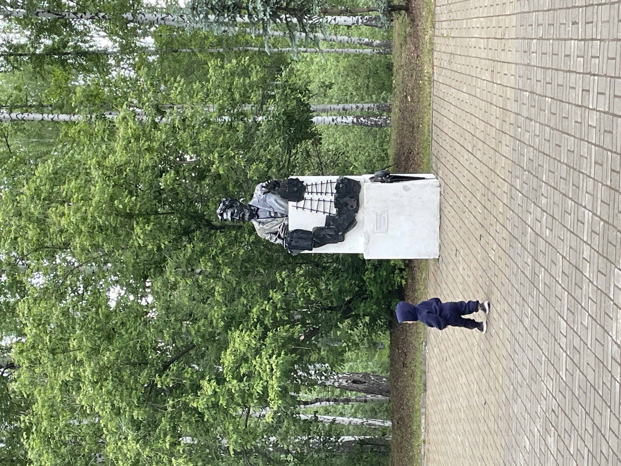 A young child wearing a dark blue hooded outfit and dark shoes stands on a light gray brick sidewalk next to a white pedestal supporting a dark gray and black sculpture. The sculpture has some grid-like elements. Behind the sculpture and pedestal is a green area with tall trees, some of which have distinctive white bark indicating birch trees. The photograph was taken in Perm, Russia.