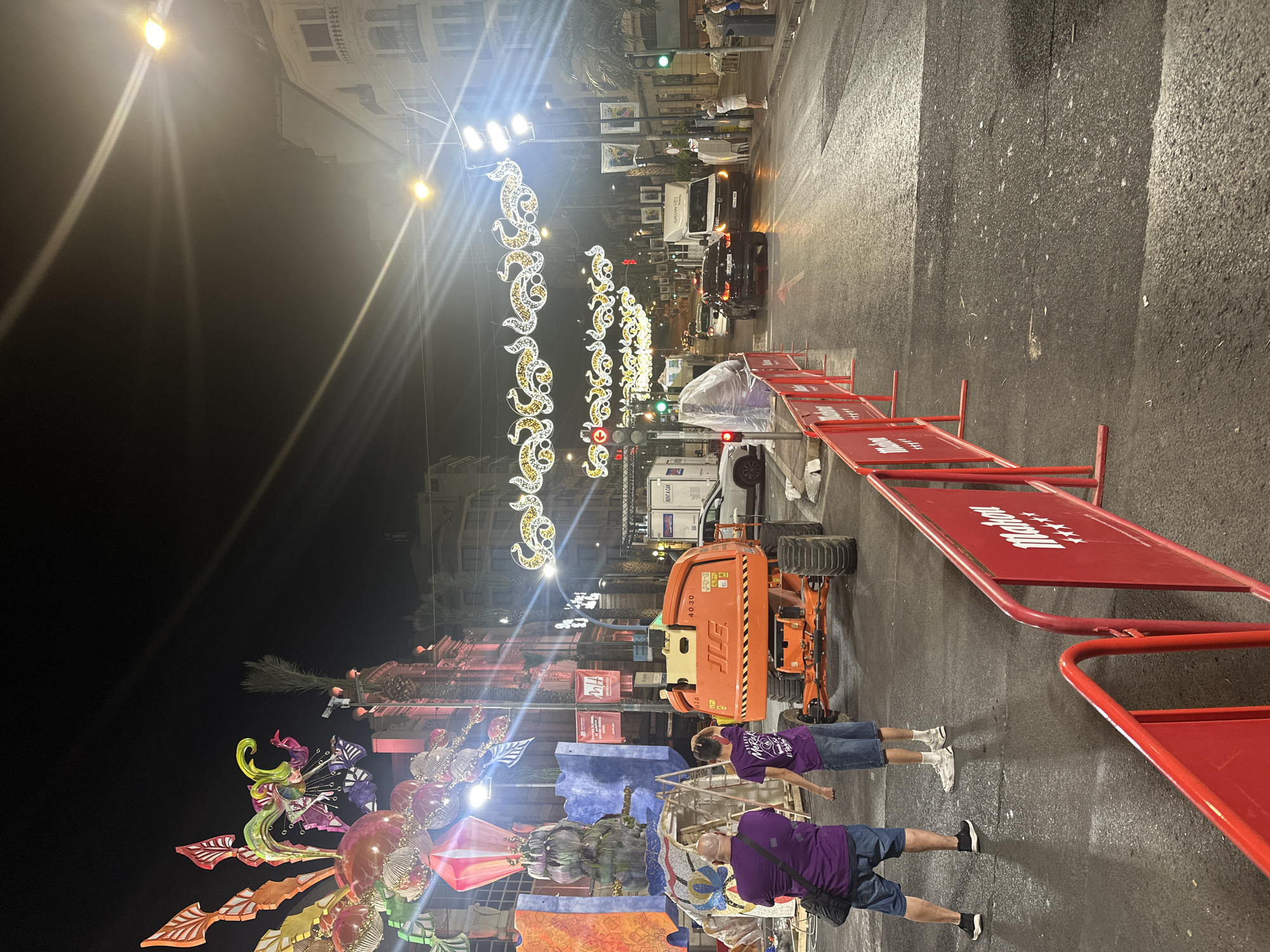The image depicts a night scene in Alicante, Spain, likely during a festival or event setup. In the foreground, two individuals, one wearing a purple t-shirt and denim shorts, and another in a purple t-shirt and jeans, stand facing a large orange scissor lift. The scissor lift, with the "JLG" logo visible, appears to be positioned on the street.
Behind the individuals, a partially constructed float or artistic installation is visible, featuring vibrant colors, spherical elements, and intricate designs, suggesting preparation for a parade or public display. The street itself is wet, reflecting the overhead streetlights and decorative illuminations that span across the road, which are elaborate and festive in nature. Buildings line the street, illuminated by artificial light. Traffic lights and street lamps are also present, indicating an urban environment.
Several vehicles, including a white van and passenger cars, are visible on the road, with some appearing to be in motion. Red barricades, some with the "Mahou" logo, are placed along the side of the street, delineating an area and likely for crowd control. The overall atmosphere suggests activity and preparation for a public event at night in Alicante.