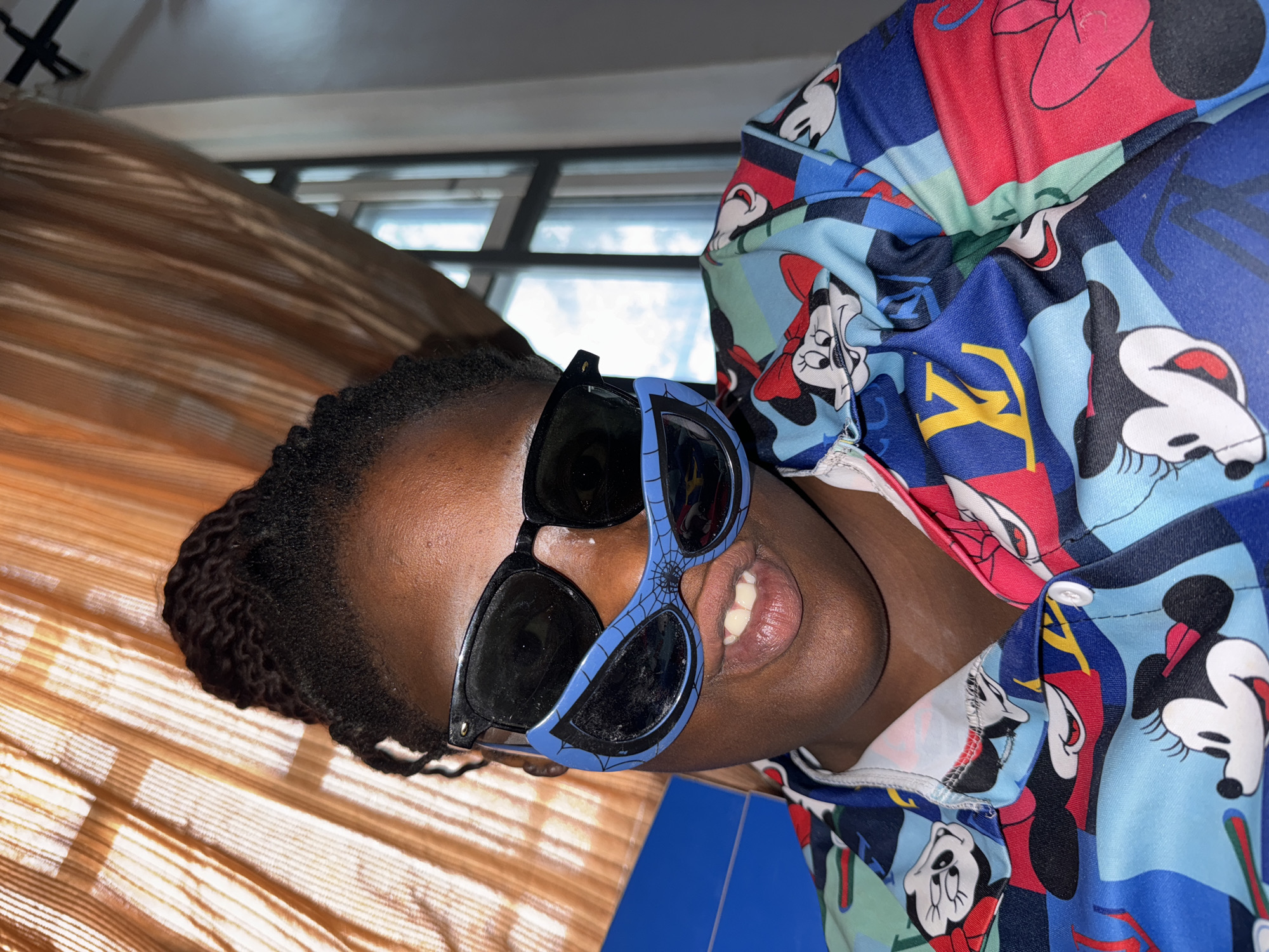 A person with dark skin is wearing two pairs of sunglasses, one black and one blue with a spiderweb design, and a colorful button-down shirt with Mickey Mouse and Minnie Mouse designs. The person is smiling slightly, showing their teeth. The background includes window blinds and a window with daylight coming through. There is no indication that the image was taken in Sabon Tasha, Nigeria.