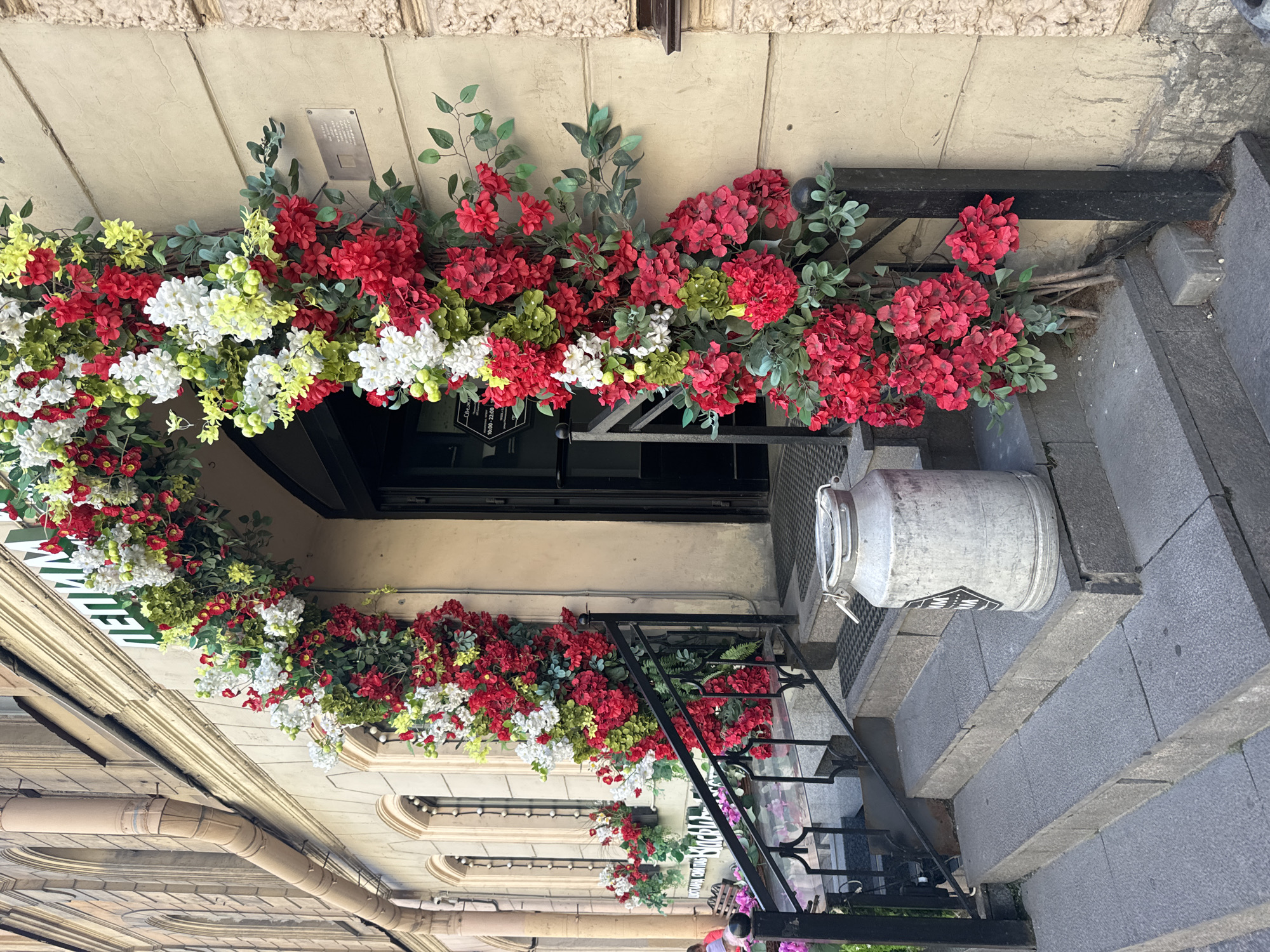 The image shows an exterior view of a building entrance decorated with a lush wreath of artificial flowers in red, white, and yellow hues, interspersed with green foliage. The flowers are arranged on a dark metal railing that frames the entrance. Below the floral arrangement, a black metal staircase with grey stone steps leads down. To the right of the stairs, a large, silver, cylindrical metal container, possibly a milk churn, rests on the steps.
The building facade is a light, creamy color with textured stone elements above the entrance. Above the flower wreath, a small rectangular plaque is visible on the wall. The entrance itself features a dark door or window. On the left side of the image, parts of the building's architectural details, including curved moldings and pipes, are visible.
A sign with green lettering is partially visible on the left, behind the flowers, displaying the word "WINE" with additional text below. Another small sign with white text on a black background is also attached to the railing near the entrance. The overall impression is that of a charming, possibly European-style establishment. The lighting suggests it is daytime, and the scene appears to be outdoors on a street or sidewalk. There are no people prominently featured in the image, except for a small glimpse of someone wearing a red shirt and another individual in the bottom right corner. The setting appears to be Saint Petersburg, Russia, as indicated by the location context.