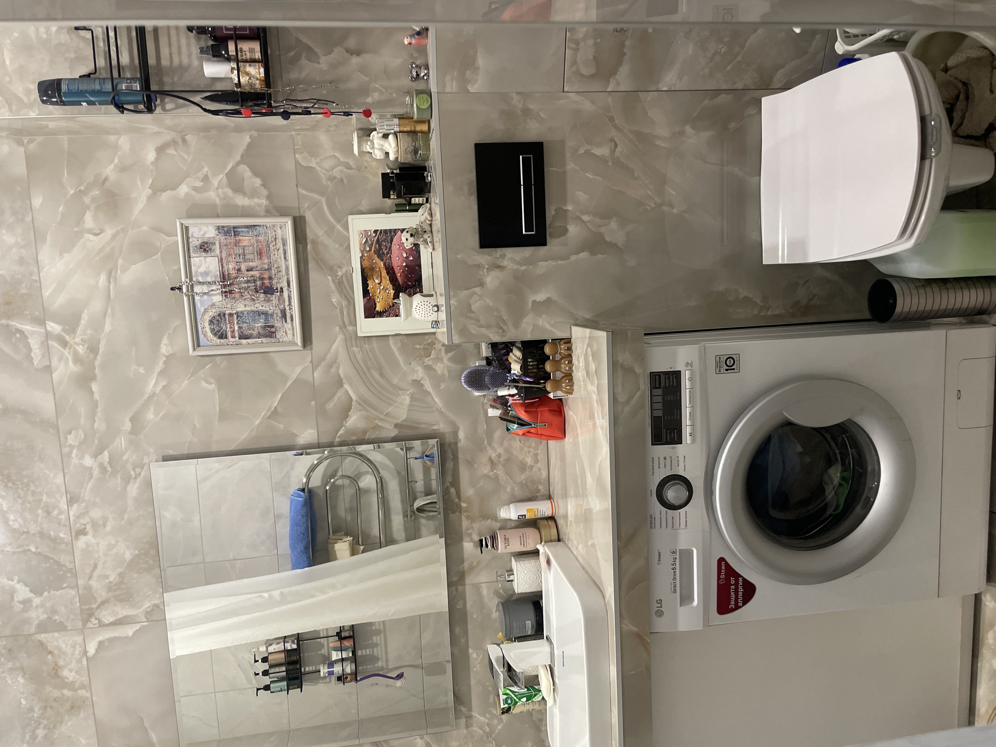 The image depicts a bathroom interior. A white washing machine is positioned next to a toilet. Above the sink are various toiletries and cosmetic items. The walls are covered in marble-patterned tiles. Framed artwork is displayed on the walls. The location appears to be Krasnoyarsk, Russia, as indicated by the text "Защита от аллергии" on the washing machine, written in Russian. There are no people visible.
