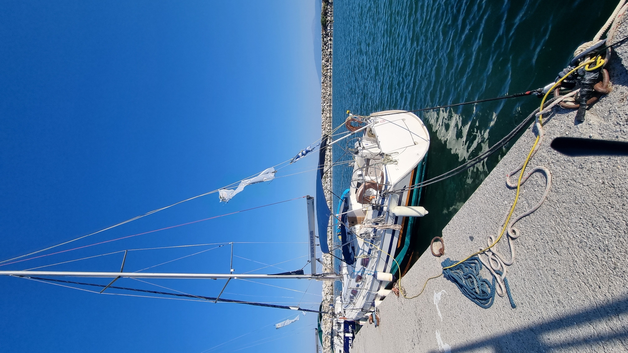 The image depicts a sailboat docked at a pier in Limani Litochorou, Greece. The sailboat, a Bavaria Cruiser, is white with teal and orange accents along its waterline. It is moored with thick ropes to the concrete pier. The boat has a tall mast with several lines running to the deck, and a blue canopy is extended over the cockpit. Two white fenders hang from the port side of the hull. Several white flags are attached to the mast, two of which appear tattered and are flapping in the breeze. A second sailboat is visible further down the pier.
The pier is constructed of concrete and shows signs of wear. Various ropes, including a coiled blue rope and a yellow rope, are scattered on the pier's surface. A rusty metal ring is visible near the coiled blue rope. The water is a clear, deep blue-green and appears calm with small ripples.
In the background, a breakwater made of large stones separates the harbor from the open sea. Beyond the breakwater, a low, hilly coastline is visible under a bright, clear blue sky. The sun casts shadows on the pier, indicating it is daytime. No people are visible in the image.
