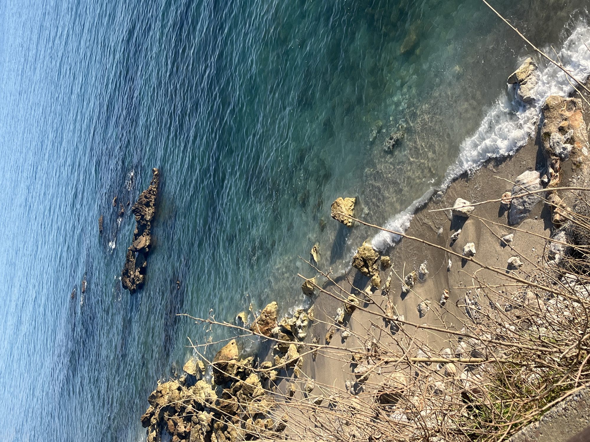 This overhead shot captures a serene coastal scene in Alanya, Turkey, from a high vantage point, likely a cliff or elevated pathway. The image is oriented vertically, looking down towards the water and a narrow stretch of shore.
The dominant subject is the sea, displaying a beautiful gradient of blues and greens. Further from the shore, the water is a deep, tranquil blue, suggesting greater depth. Closer to the land, it transitions to a clearer, vibrant turquoise, revealing the rocky seabed and scattered pebbles beneath the surface. Small, rippling waves are visible across the water's surface, reflecting sunlight.
Along the shore, gentle white waves crash against a mix of golden-brown sand and numerous rocks, creating patches of sea foam. Large, irregular rocks of various sizes are scattered both in the shallow water and along the sandy strip, some appearing rugged and dark, others lighter and smoother.
In the immediate foreground, a dense network of thin, bare, dry branches and twigs extends into the frame from the right and bottom, partially obscuring the view of the coastline directly below. A hint of green foliage is visible among the twigs at the very bottom right.
The scene depicts a calm, sunny day, with clear weather. No people or human structures are visible, emphasizing the natural beauty of the rocky coastline and the clear waters. There is no visible text in the image.