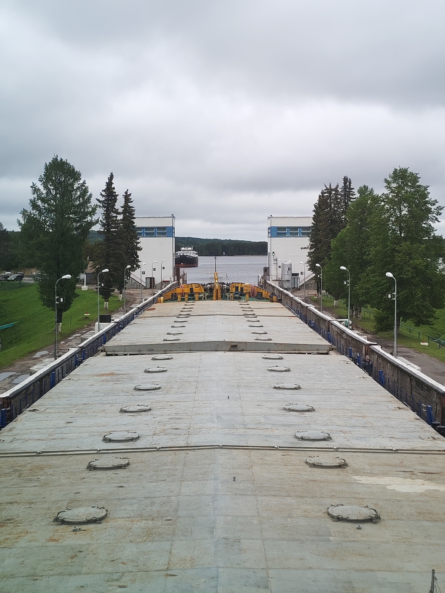The image depicts a section of the Volga-Baltic Waterway located in Белоусово, Russia. A large, flat, concrete or metal structure with numerous circular hatches dominates the foreground, extending into the distance. This is likely part of a canal lock or dam system. In the background, a waterway is framed by two identical, white, rectangular buildings with blue trim. A ship is partially visible on the waterway, between the buildings. The sky is overcast. Trees line the edges of the canal, adding to the scenery. Streetlights are visible along the side.