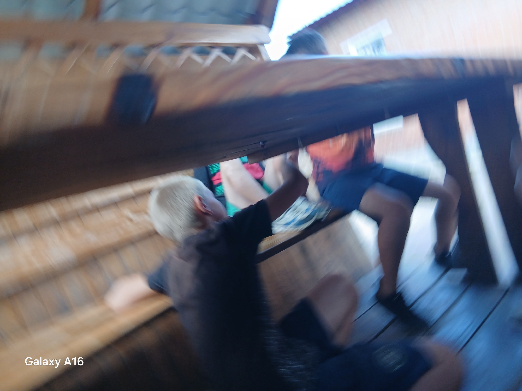 The image depicts an outdoor scene, likely a wooden structure with a slatted roof and wooden floorboards, suggestive of a picnic area or gazebo. There are at least three individuals partially visible and blurred due to motion. In the foreground, a person with blonde hair is seen from the side, wearing a dark t-shirt and black shorts, appearing to be in motion. Another person, wearing an orange and black patterned shirt and dark shorts, is visible in the mid-ground, also in motion. Legs and feet of other individuals are also discernible on the wooden floor. The overall environment appears to be rustic and possibly rural. The "Galaxy A16" watermark indicates the device used to capture the image. The image is not sufficient to confirm the specific location within Ustyvytsia, Ukraine.