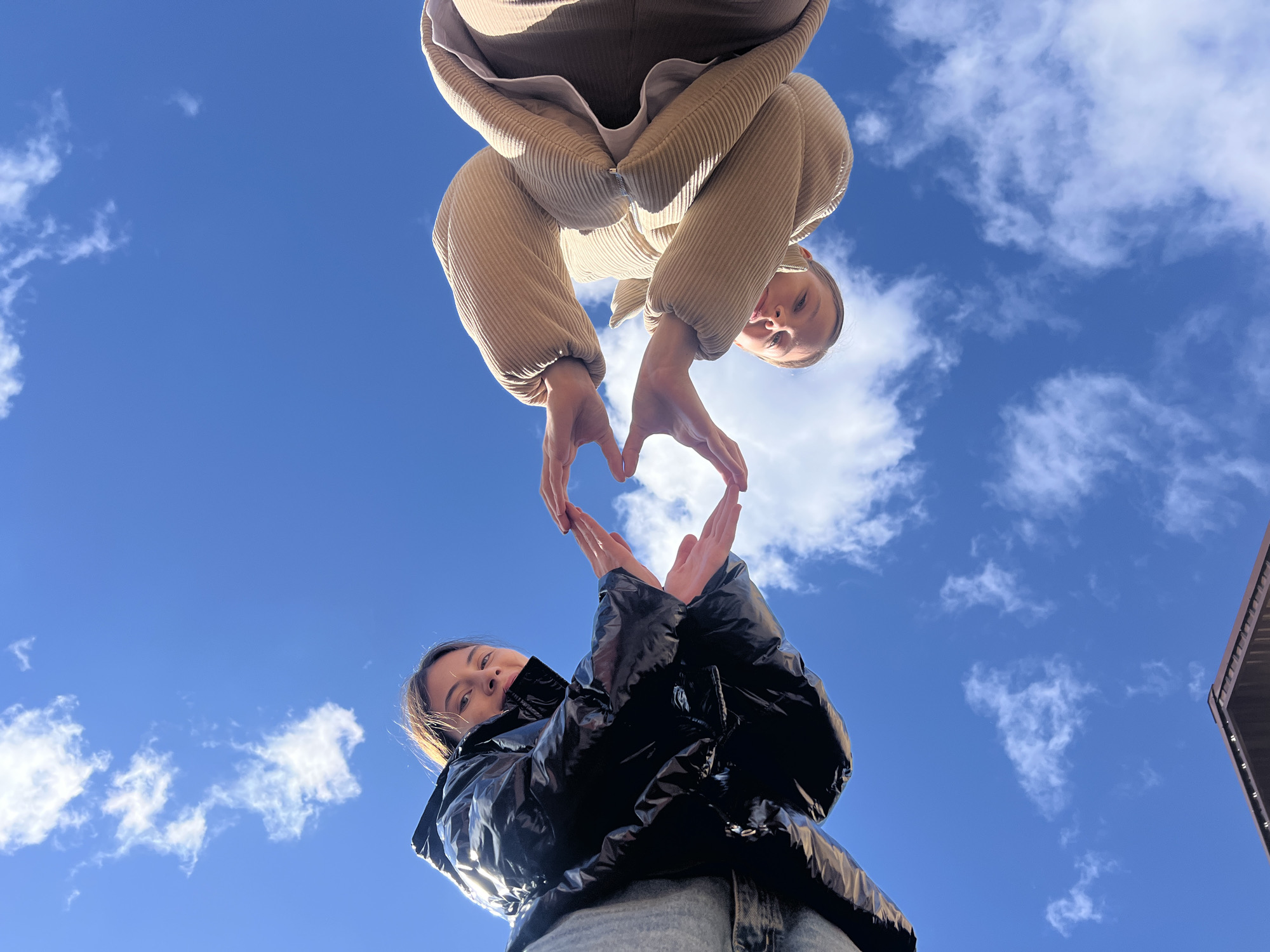 Two young women are posed in an upward-looking shot against a blue sky with scattered clouds. The woman at the top of the frame wears a light brown, corduroy-textured puffer jacket. She holds her hands to form the top half of a heart shape. The woman at the bottom of the frame wears a black, shiny puffer jacket and gray jeans. She holds her hands to complete the bottom half of the heart shape. Their faces are visible and turned toward the camera. Part of a building is visible in the upper right corner. This image could have been taken anywhere, the city of Votkinsk, Russia is not determinable from the image.