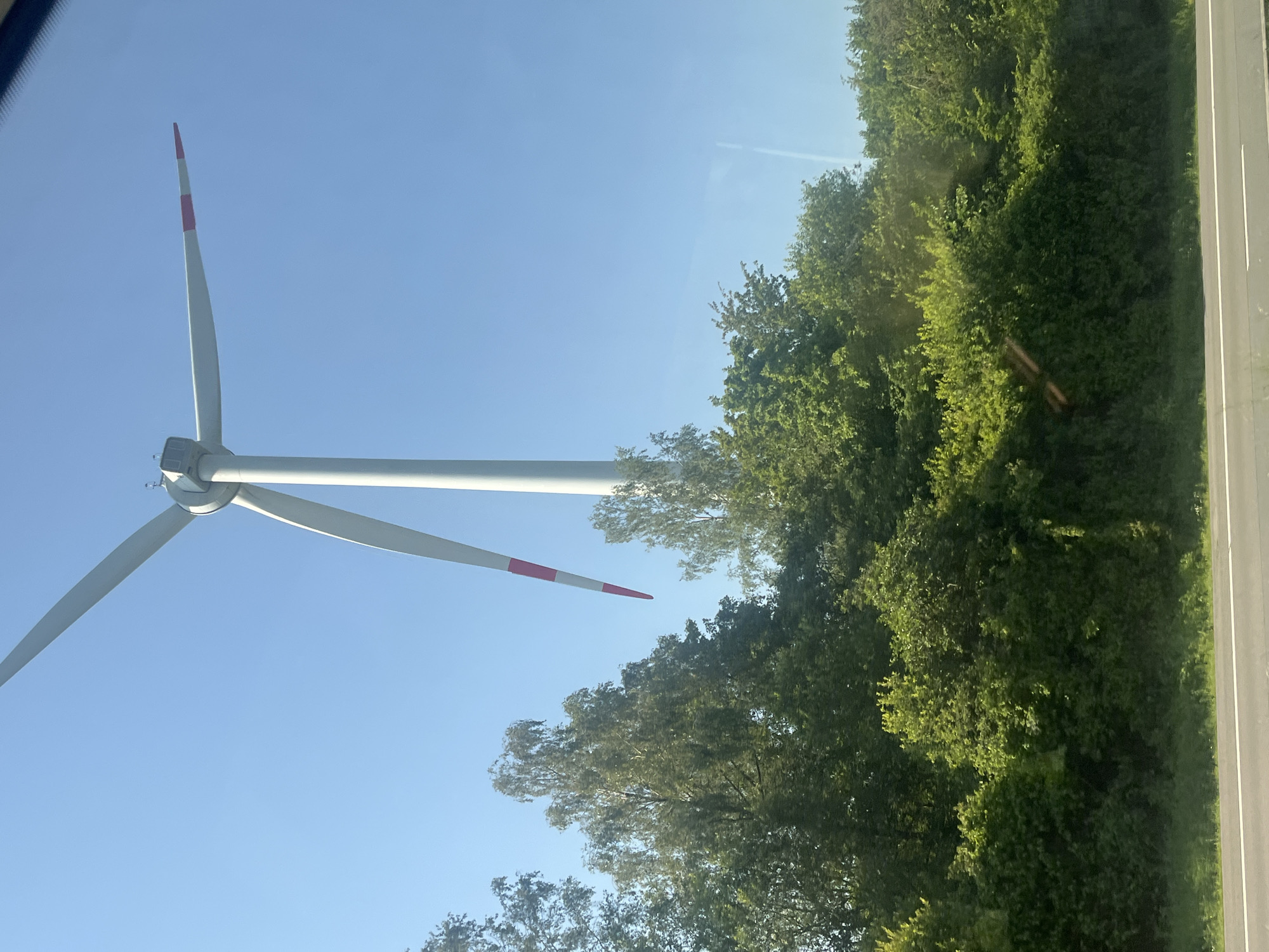 The image shows a wind turbine against a clear blue sky. The wind turbine is tall with three white blades, two of which are visible, each with red markings near the tips. In the foreground, there is a dense line of green trees and bushes, suggesting a natural or rural setting. Below the trees, a grey road with white lines is visible, indicating it might be a highway or main road. The scene appears to be captured from a moving vehicle due to the slight blur and the angle of the shot. The image does not depict Courcelles, Belgium, nor does it show any people or specific actions. It is a landscape photograph of a wind turbine and surrounding vegetation.