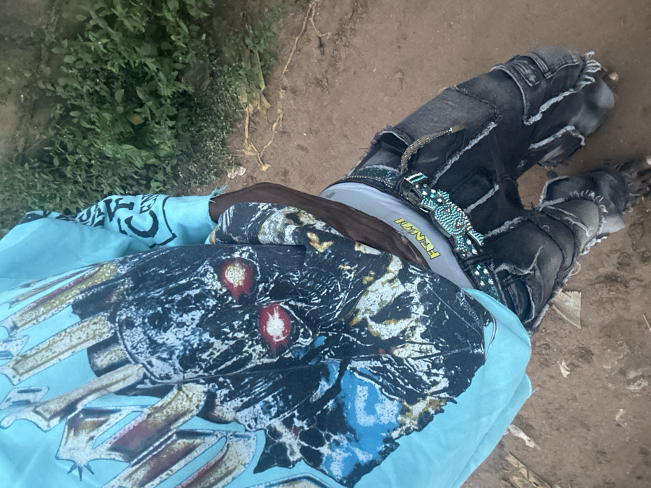 A person, seen from above, wears tattered dark denim jeans with a sparkly belt and designer-labeled underwear. A t-shirt with a skull-like graphic in blue and white obscures the upper torso. To the left, green foliage is visible, while the ground to the right is dusty brown. The overall image presents a casual, edgy style in an outdoor setting.