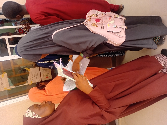 The scene is set inside a public service or registration office in Kwazailewa, Nigeria. Six individuals are present. In the foreground, a young female with light brown skin, wearing a maroon hijab and dress with a patterned forehead band, holds an open document or book in her left hand and a pink-cased smartphone in her right. She directs her gaze towards the right. To her right, another young female, partially viewed from behind, wears a dark grey hijab and dress, with a pink backpack adorned with multiple charms slung over her right shoulder. Her right hand is extended towards the documents held by the first female, suggesting an interaction or exchange. Both foreground females wear flat footwear; the one in grey has visible patterned anklewear.
Behind a white-framed glass partition, three individuals are visible. One person, largely obscured, wears an orange head covering and holds a phone to their ear. To their right, a young male in a red long-sleeved shirt is partially visible. Further right, another male in a patterned shirt (red, white, black) is visible through the glass. A white rectangular sign is taped to the glass partition, displaying "VOTER PLASTIC ID CARD IS AVAILABLE" and the number "8067503370", along with a small red, green, and white segmented graphic. The background includes a light-colored wall.