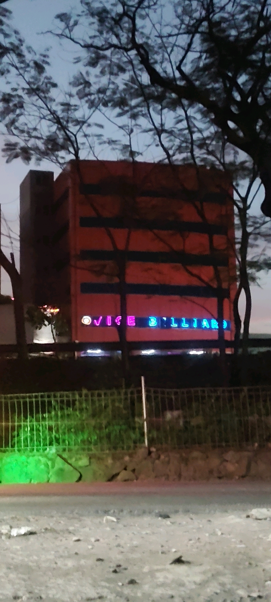 The image shows a building with red and blue neon lights that spell out "VICE BILLIARD" in Sukaresmi, Indonesia. The building is tall and has a rectangular shape. Trees with bare branches partially obscure the building and the neon sign. In the foreground, there is a metal fence, and behind it, a wall with some greenery and dirt. The sky is visible in the background and appears to be dusk or early evening. The road in front of the fence is visible, and it appears to be unpaved. There are no people visible in the image.