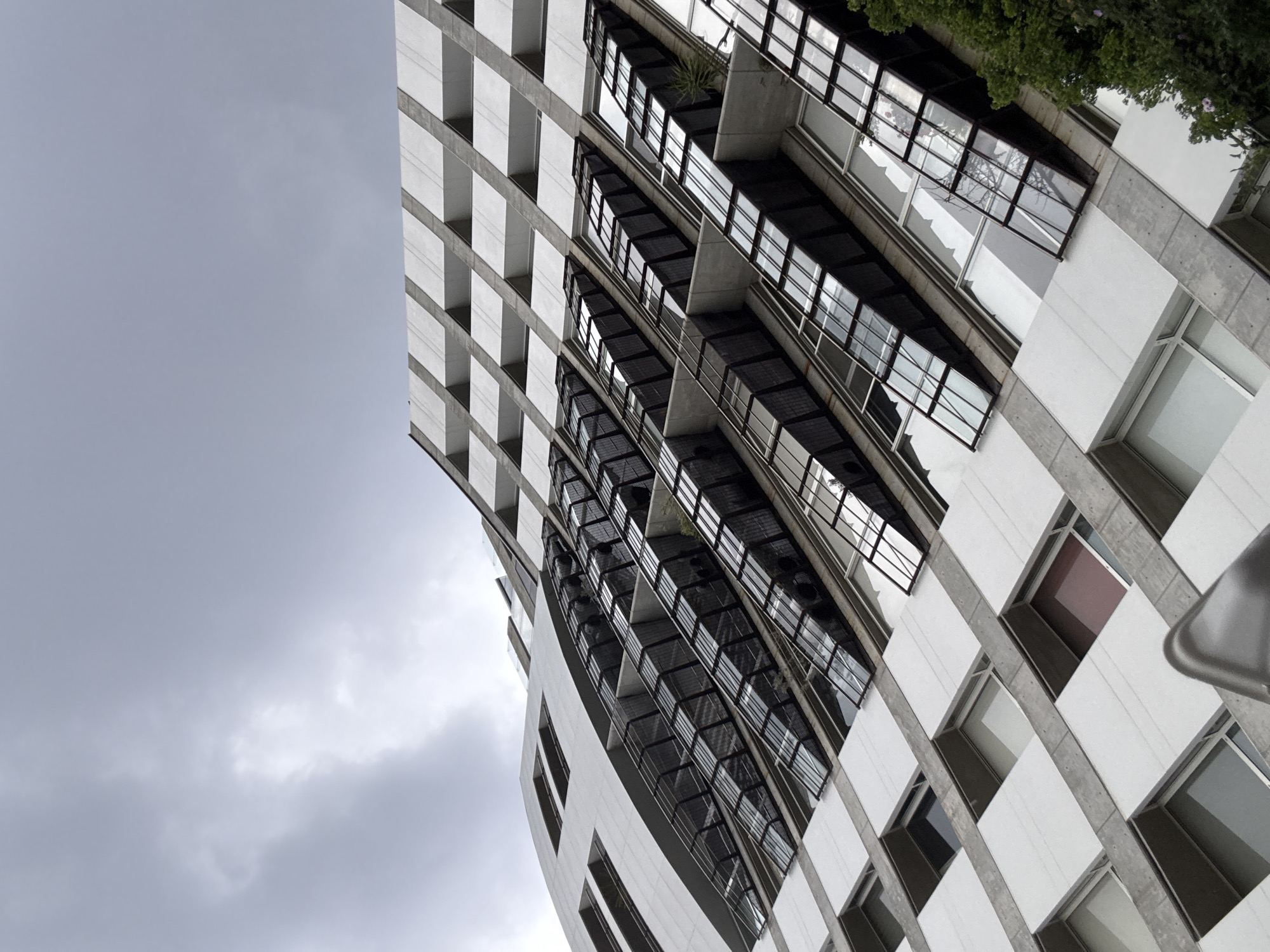 A low-angle, rotated shot captures a modern building in Puebla, Mexico, under a cloudy sky. The building's facade features a grid-like pattern of white blocks and concrete supports. A curved section of the building is made up of glass windows and balconies. Green foliage grows on some of the balconies. The gray sky is visible to the left and around the edges of the building.