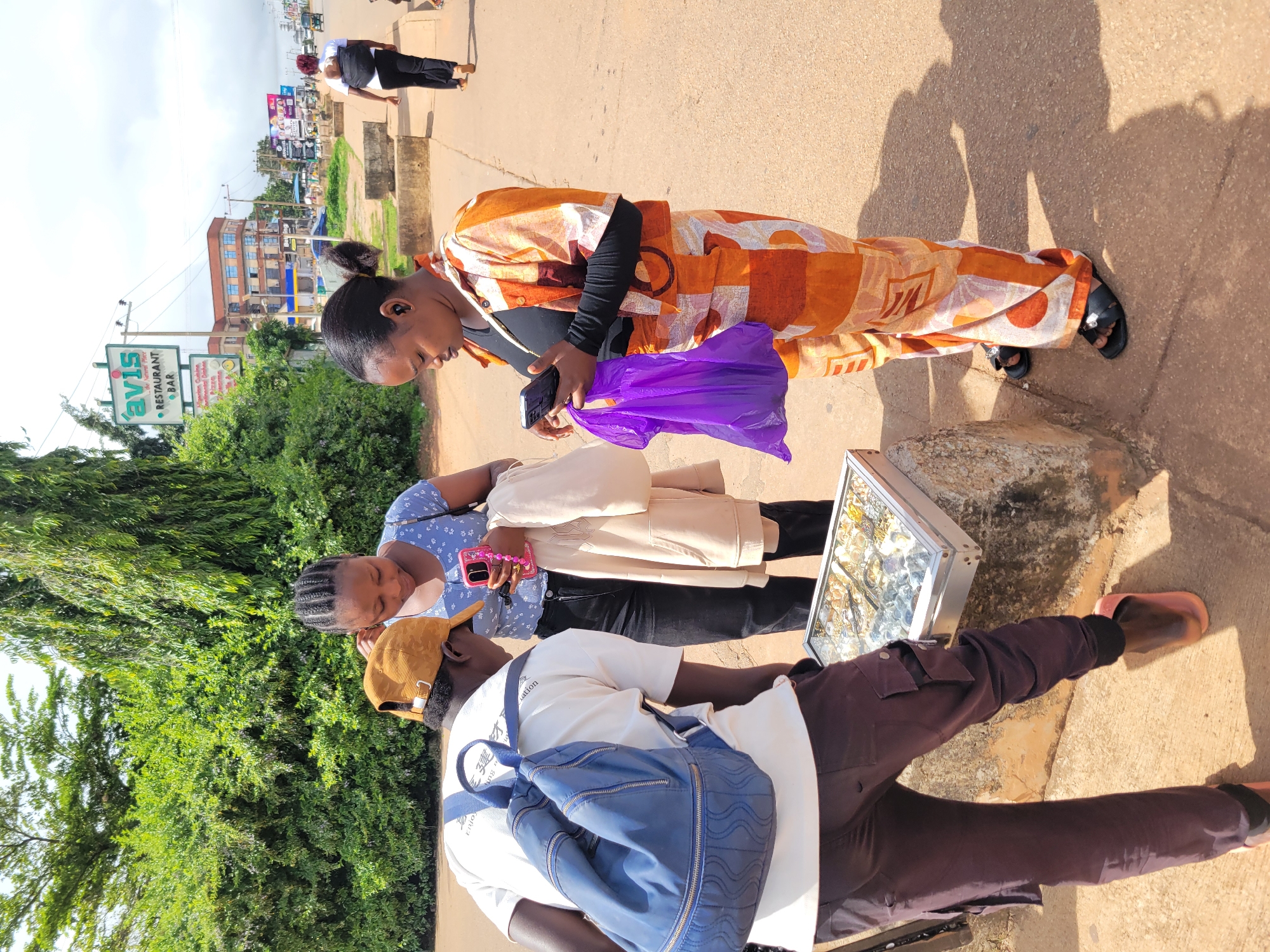 In Jos, Nigeria, three young adults are gathered around a display of jewelry on a sidewalk. A person wearing a tan baseball cap and a white t-shirt with a blue backpack is leaning over the display, examining the items. To their right, a young woman in a blue patterned top holds a pink smartphone. Another young woman, dressed in a vibrant orange and white patterned outfit, stands to the right of the jewelry, looking at her own smartphone and holding a purple plastic bag. The background reveals a street scene with a building advertising "avis RESTAURANT BAR" and other businesses, along with a pedestrian walkway and a road with vehicles. The scene suggests a casual street-side transaction or browsing activity.