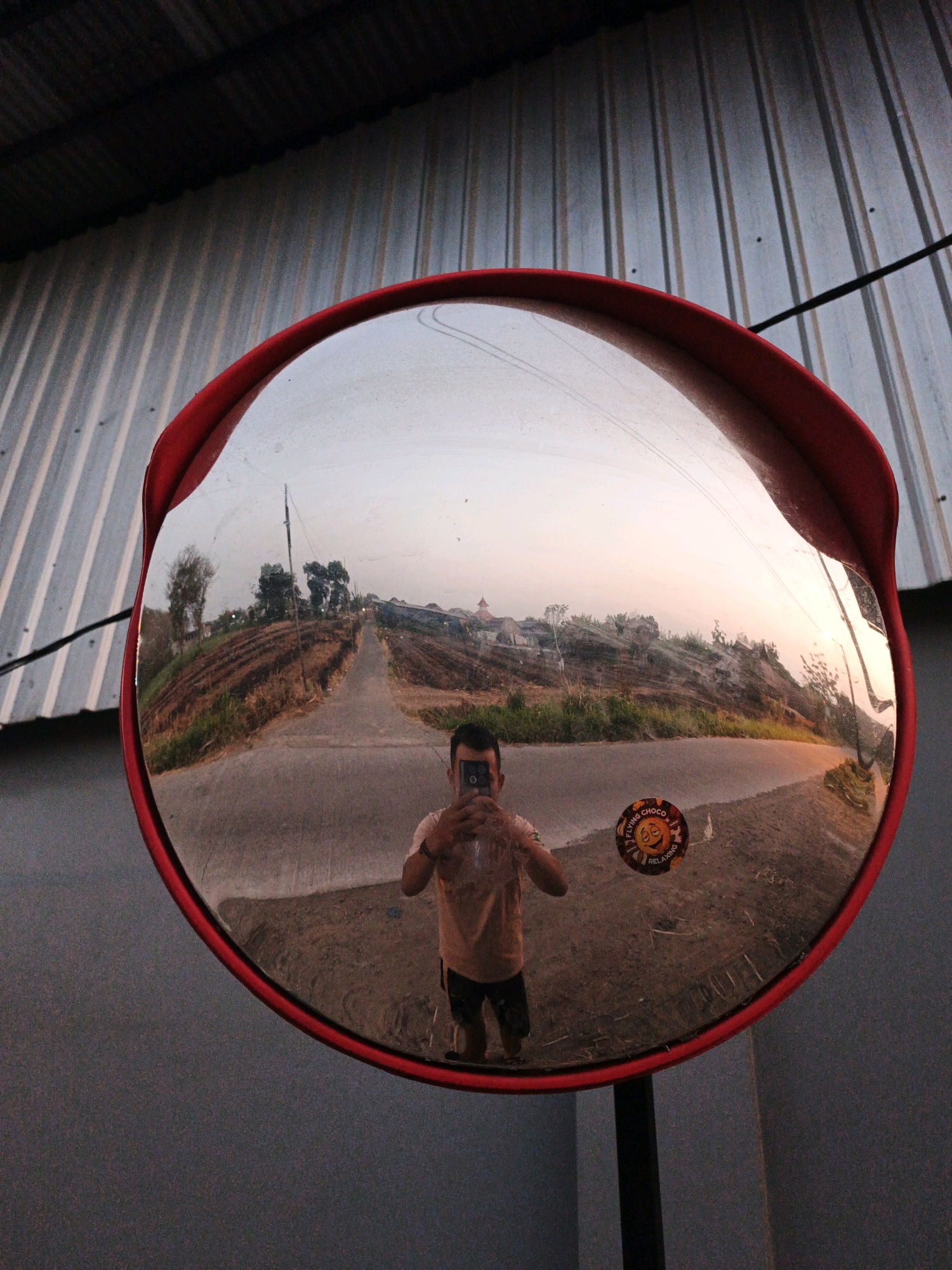 In Trowulan, Indonesia, a convex mirror with a red rim reflects a selfie taken by a person holding a smartphone. The mirror is mounted on a black pole and is positioned in front of a corrugated metal wall. The reflection in the mirror shows a road leading uphill through a rural landscape with brown, tilled fields and scattered trees. The sky is clear and shows the soft light of either sunrise or sunset. A small, circular sticker with a smiling sun graphic and text reading "FLYING CHOCO RELAXING" is visible on the lower right portion of the mirror. The person in the selfie is wearing a light-colored t-shirt and dark shorts.