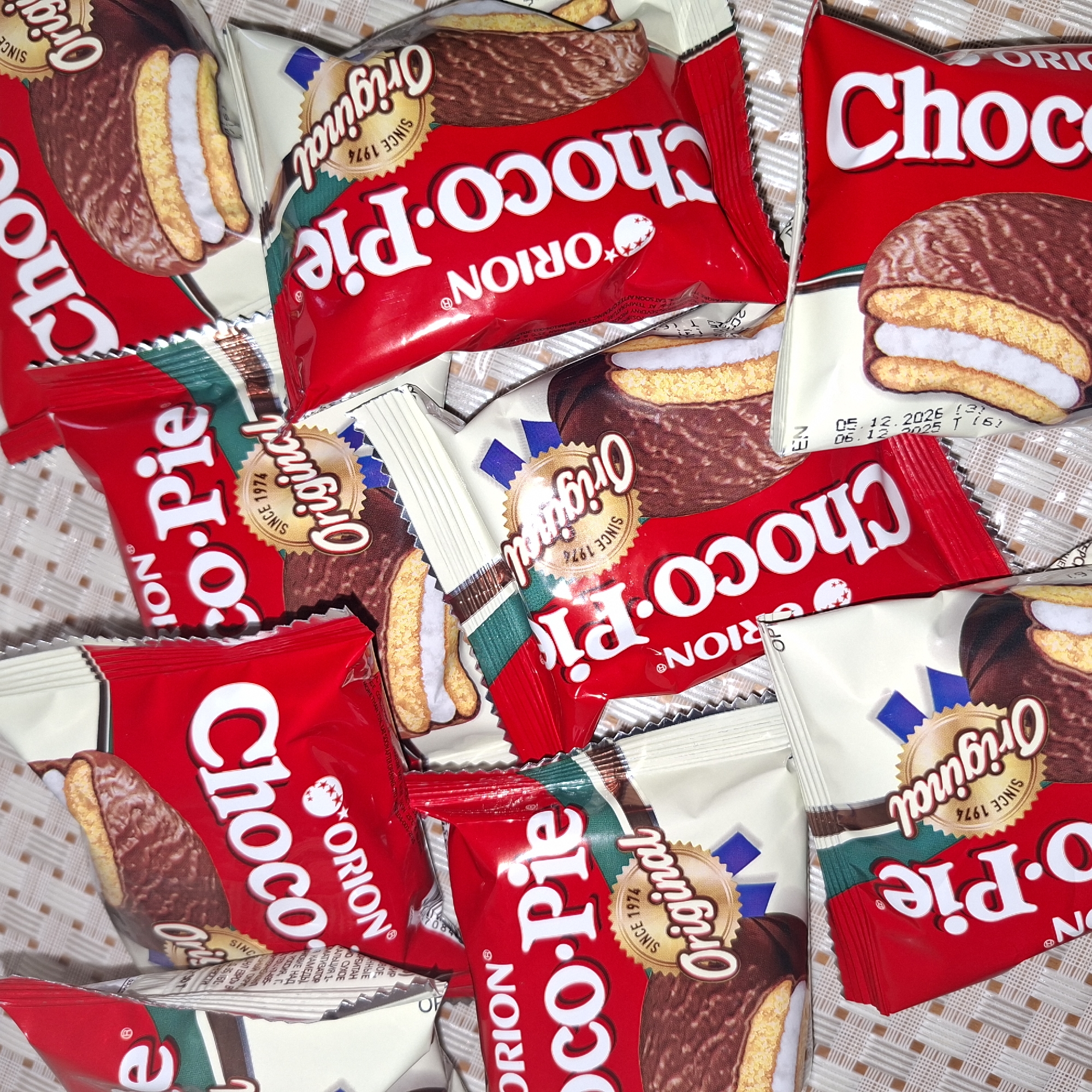 This is a close-up, overhead shot of several Orion Choco-Pie packages. The packages are predominantly red with white lettering and feature an image of a Choco-Pie cookie, which consists of two cake layers sandwiching a marshmallow filling and coated in chocolate. Each package also displays a circular "Original Since 1974" seal. The Choco-Pies are arranged in a pile, some overlapping each other, creating a dynamic, layered composition. The background appears to be a textured surface, possibly a tablecloth, with a checkered pattern in light brown and white. Some packages show visible expiry dates, such as "05.12.2026" and "06.12.2025". The lighting suggests an indoor setting.